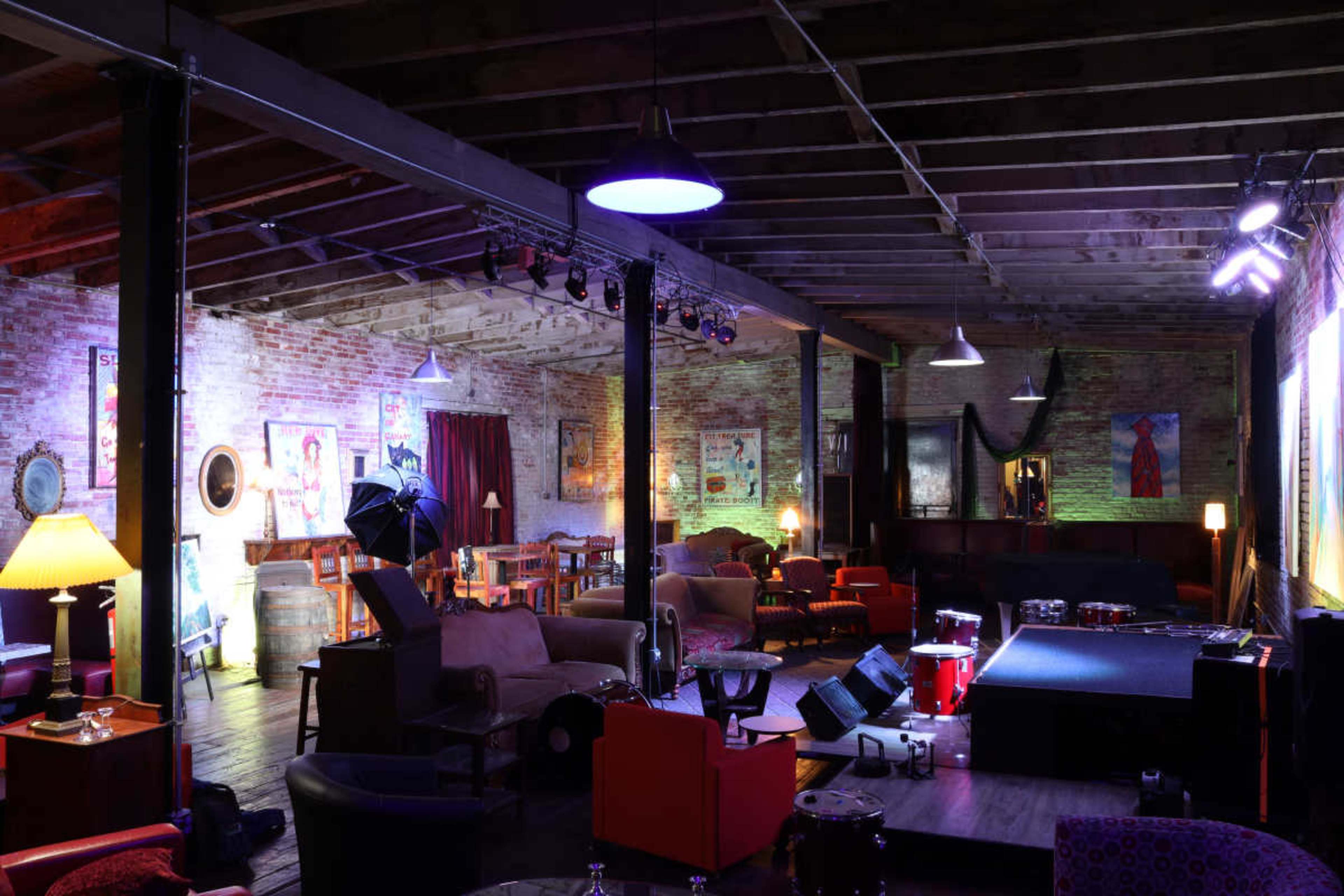 The image shows a dimly lit music or performance venue featuring exposed brick walls, various seating arrangements, and a small stage with lighting.