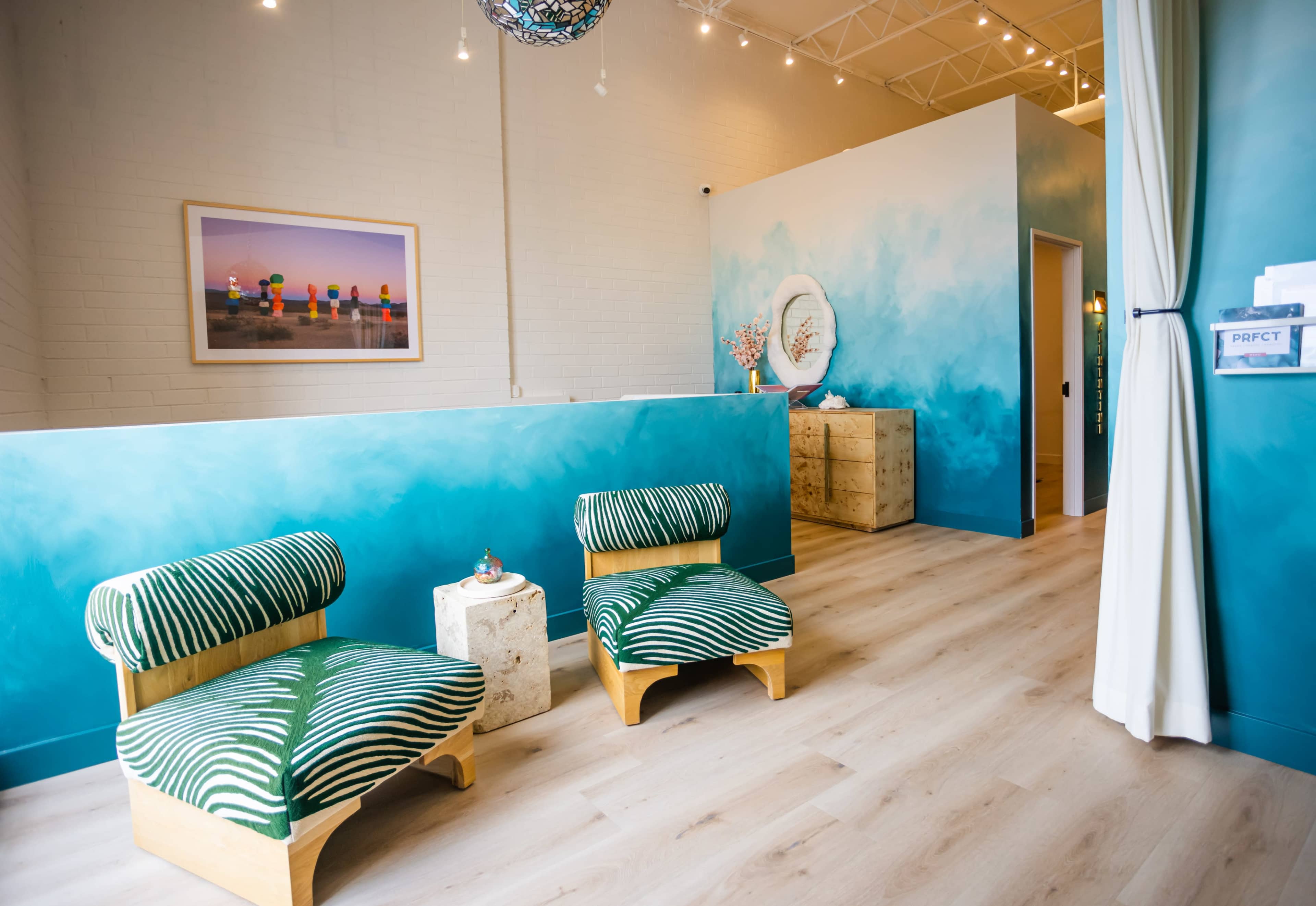 The image shows a brightly lit, modern interior of a wellness space featuring striped green chairs, a small decorative table, and a mural with blue and white hues on the walls.