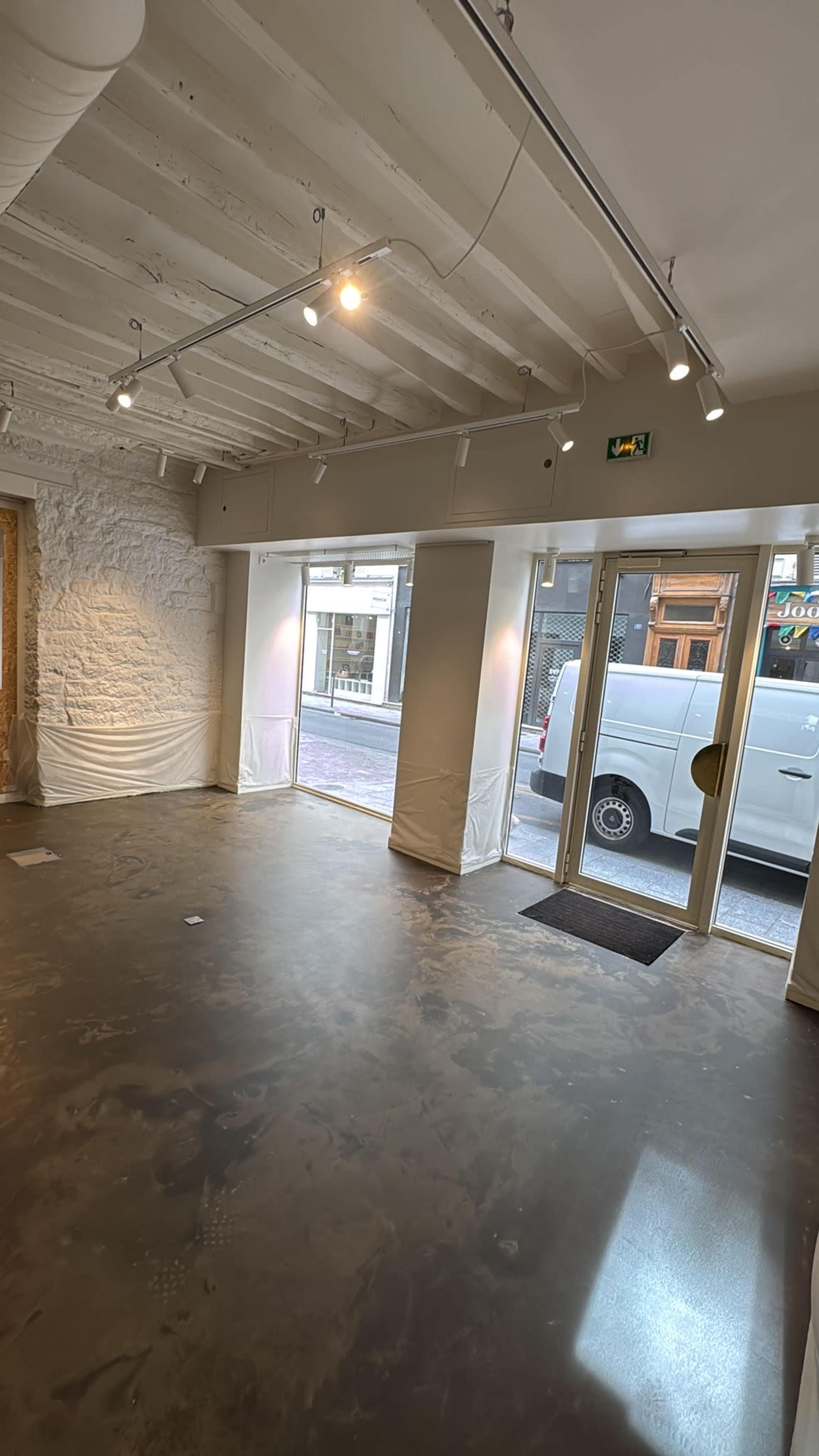 The image shows an empty interior space with white walls, a concrete floor, and large windows allowing natural light, along with a white van visible outside.