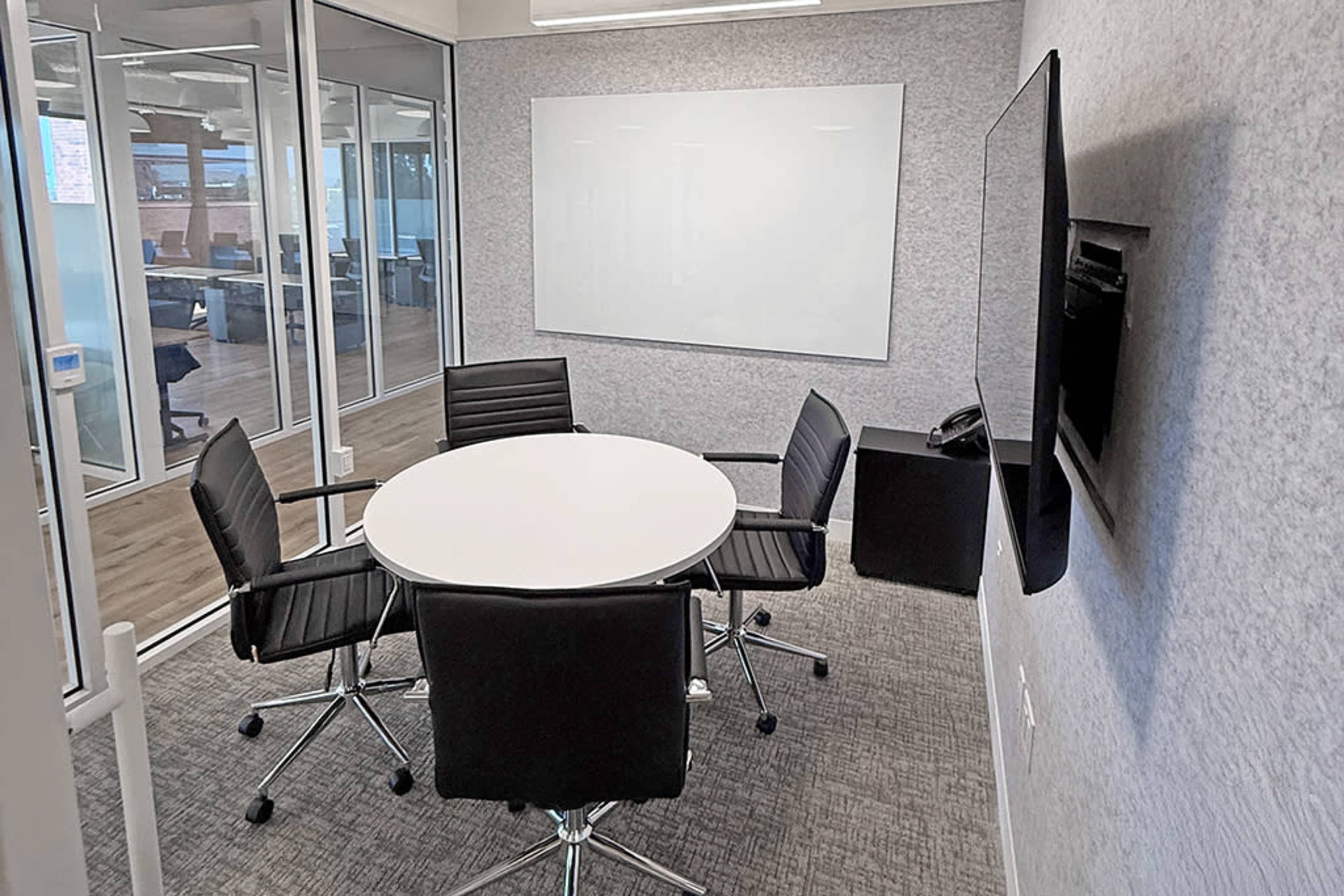 The image shows a small conference room with a round table surrounded by four black chairs, a whiteboard on the wall, and a television mounted on the opposite wall.