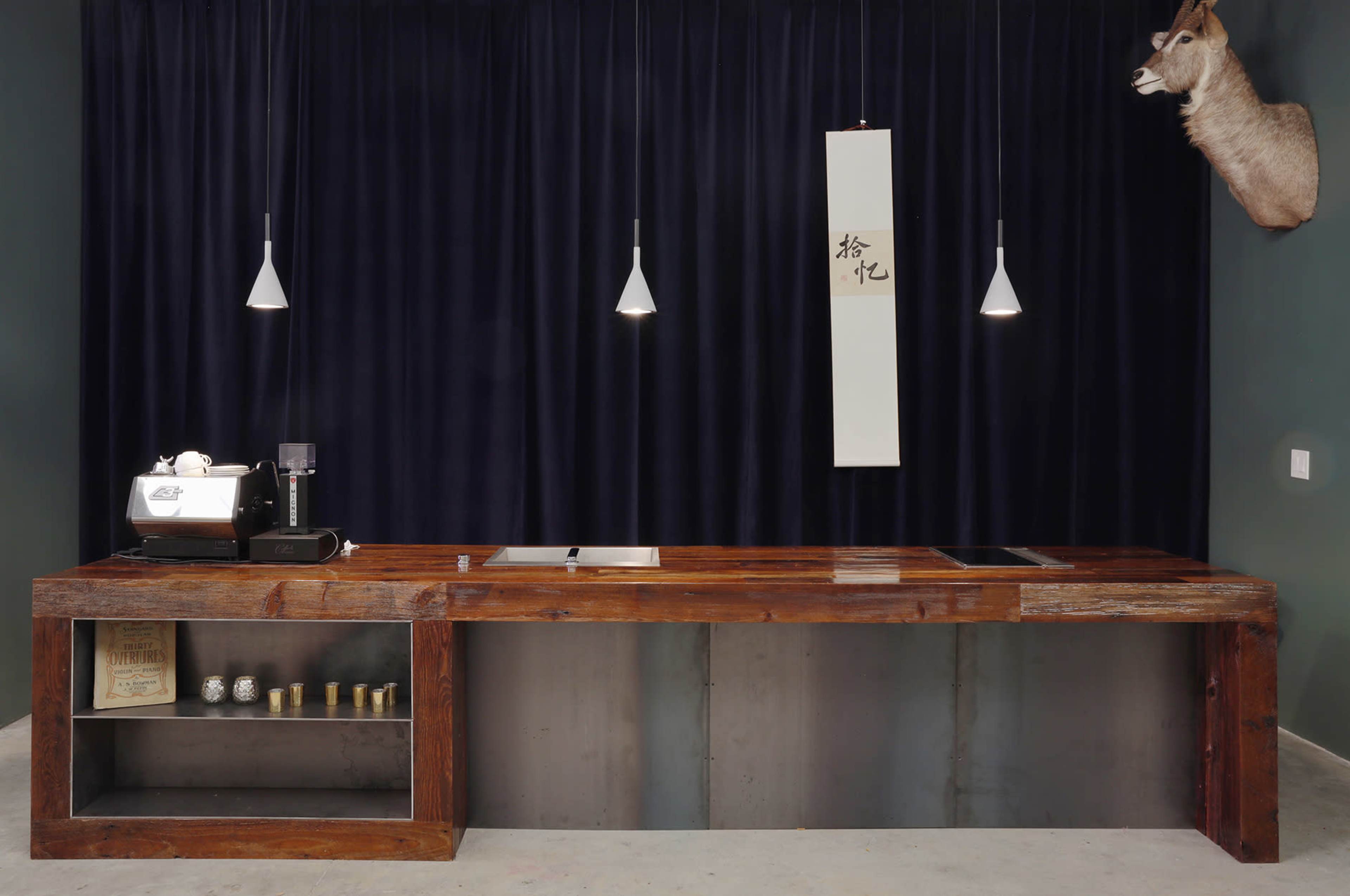 A modern kitchen countertop made of wood and metal, with a coffee machine on one side, pendant lights above, and a mounted deer head on the wall.