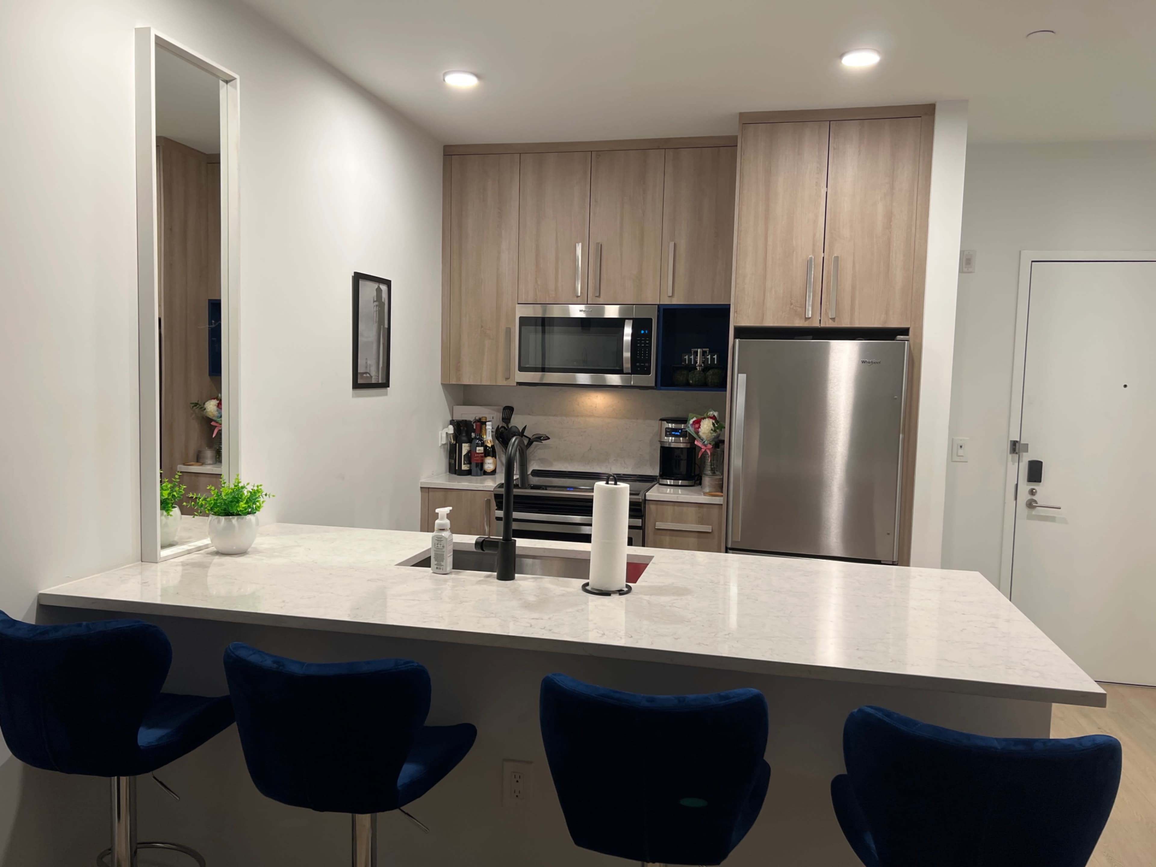 A modern kitchen with wooden cabinets, stainless steel appliances, and a white countertop with four blue bar stools.