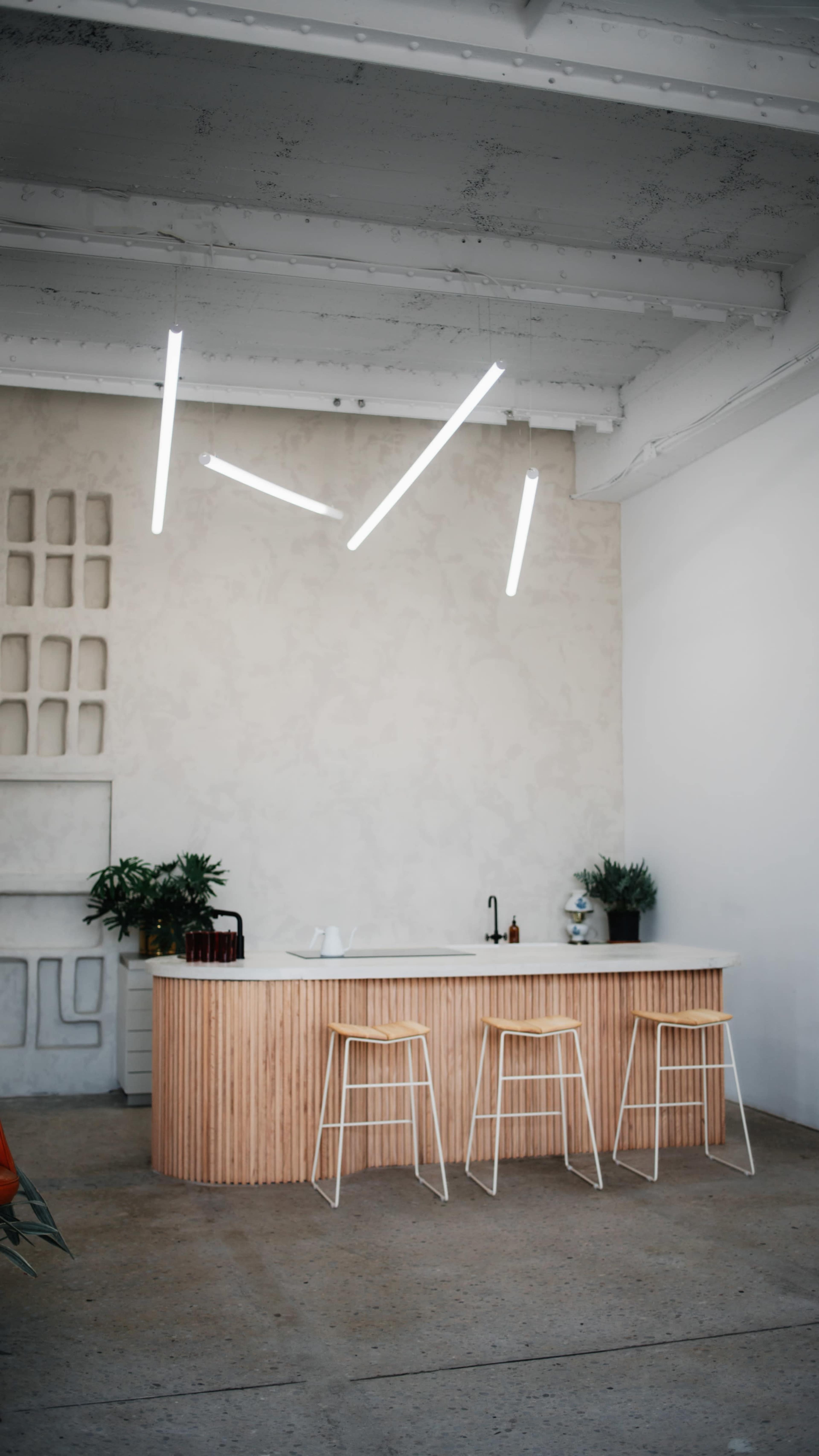 A modern kitchen features a wooden island with three stools, illuminated by minimalist overhead light fixtures, against a backdrop of a textured wall and green plants.