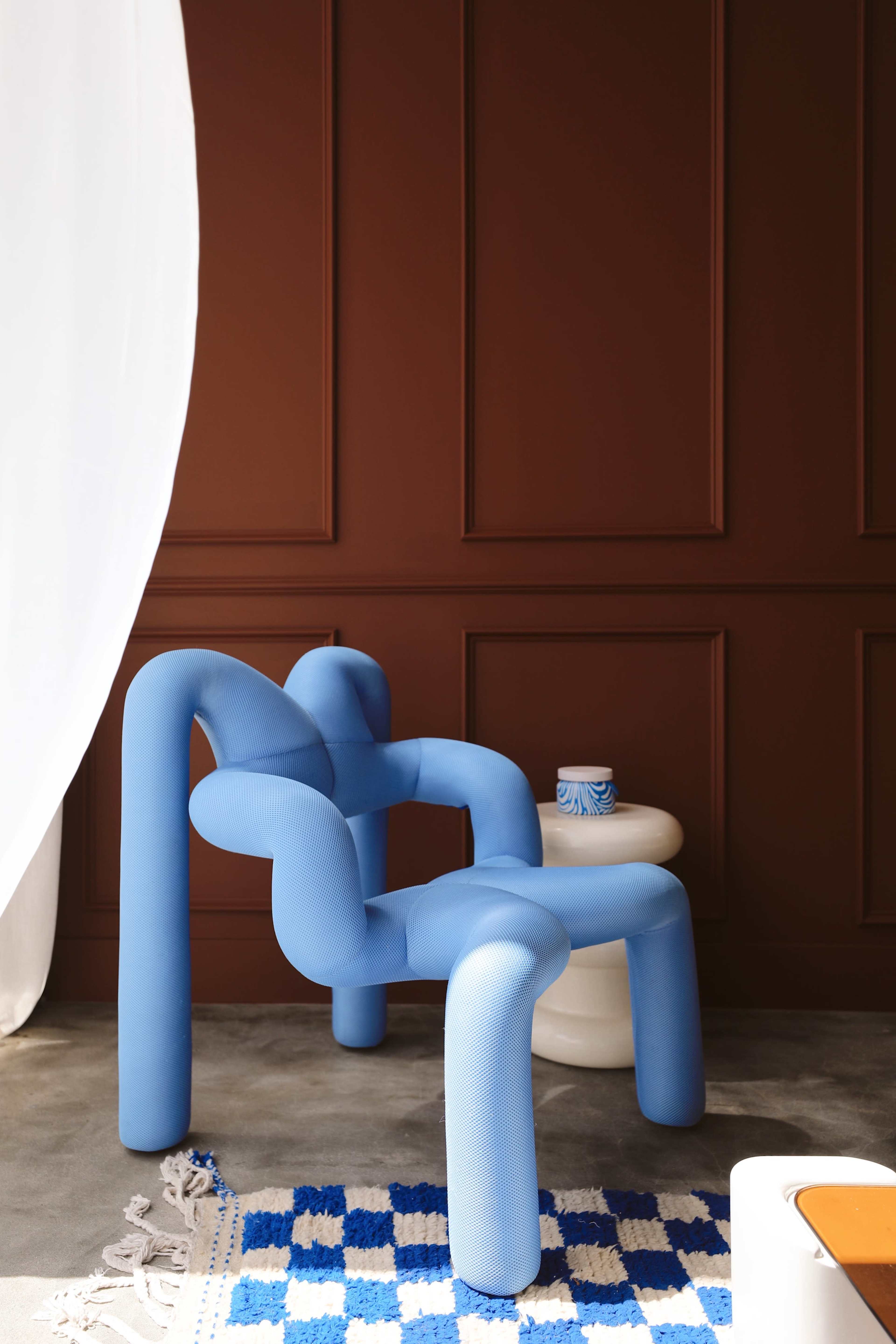 A unique blue armchair with a sculptural design next to a white side table and adorned with a patterned rug on a concrete floor.