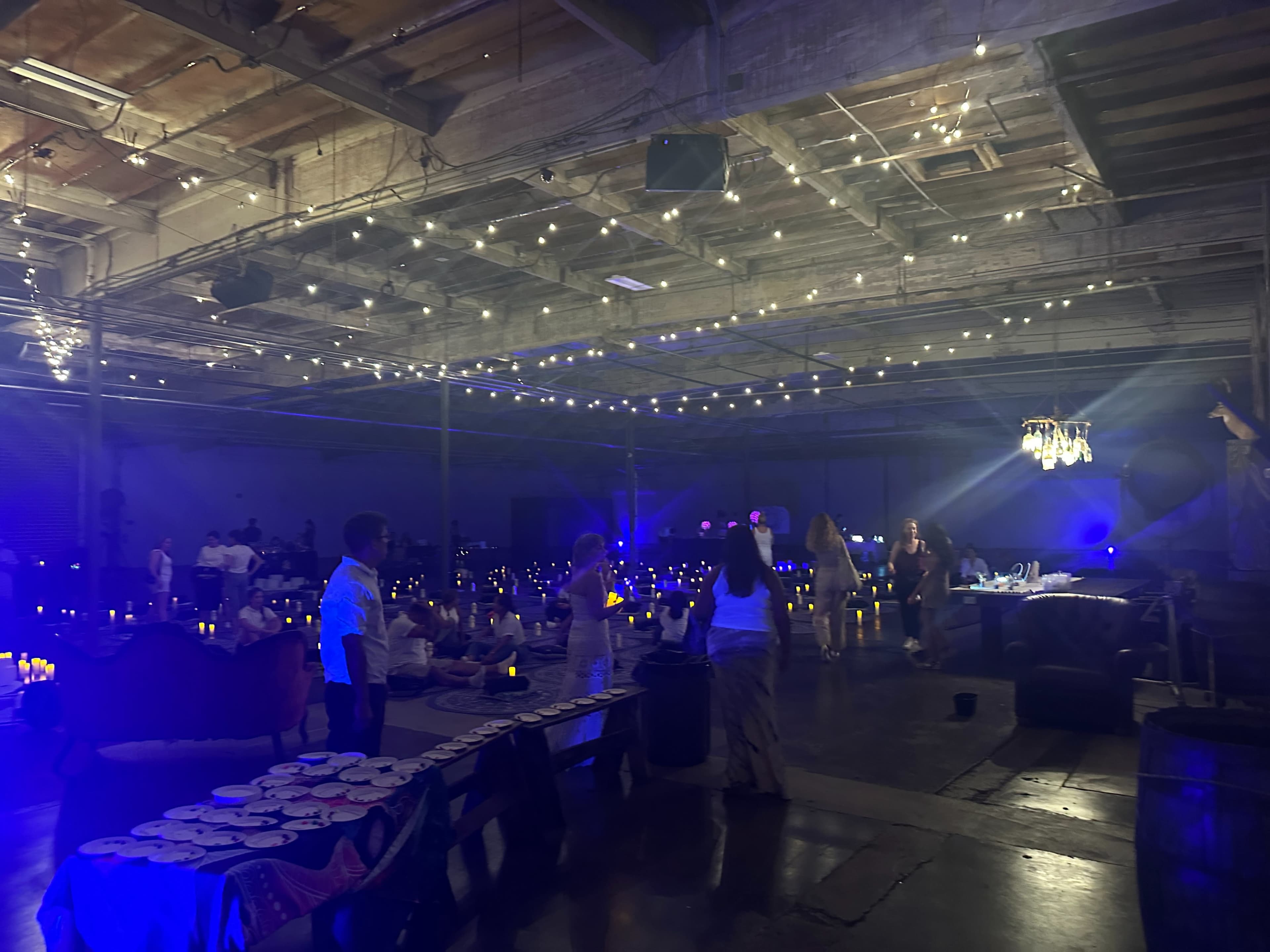 A spacious indoor venue with strings of lights overhead, set up for an event with tables arranged and guests mingling under soft blue lighting.