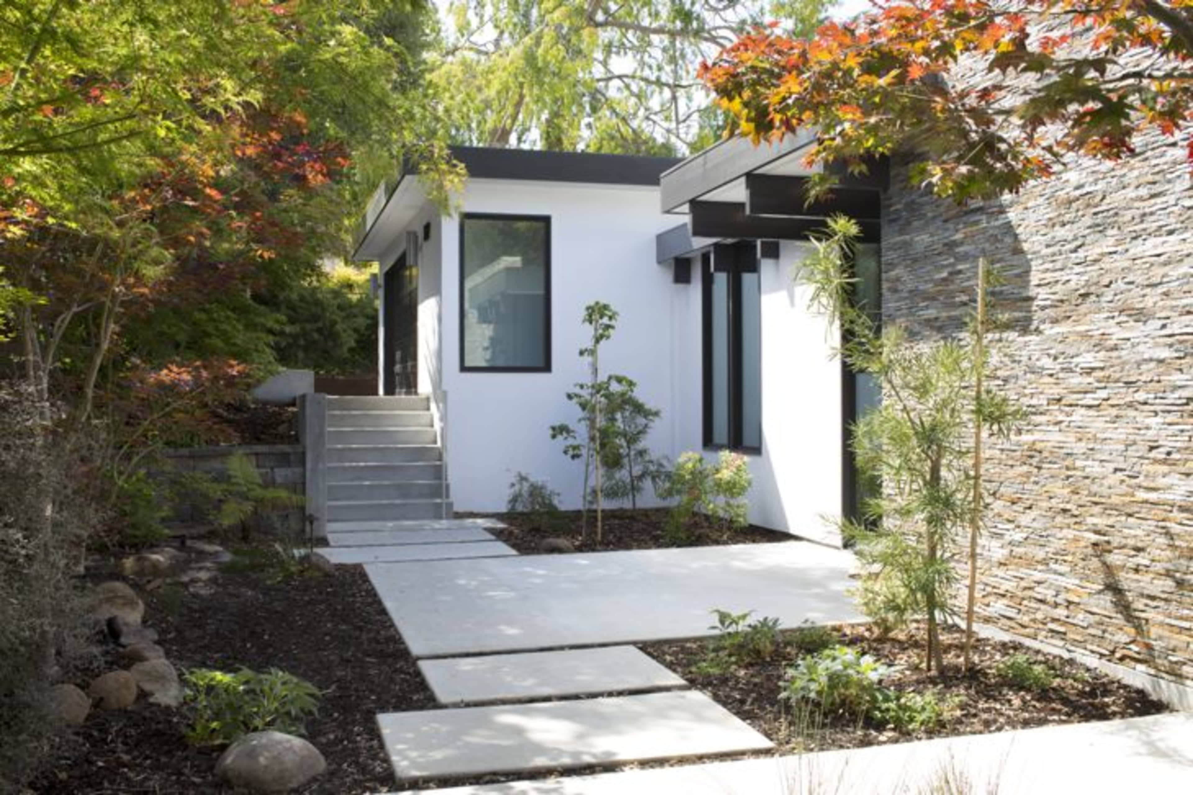 A modern home entrance with a concrete pathway, surrounded by landscaped greenery and a stone wall feature.