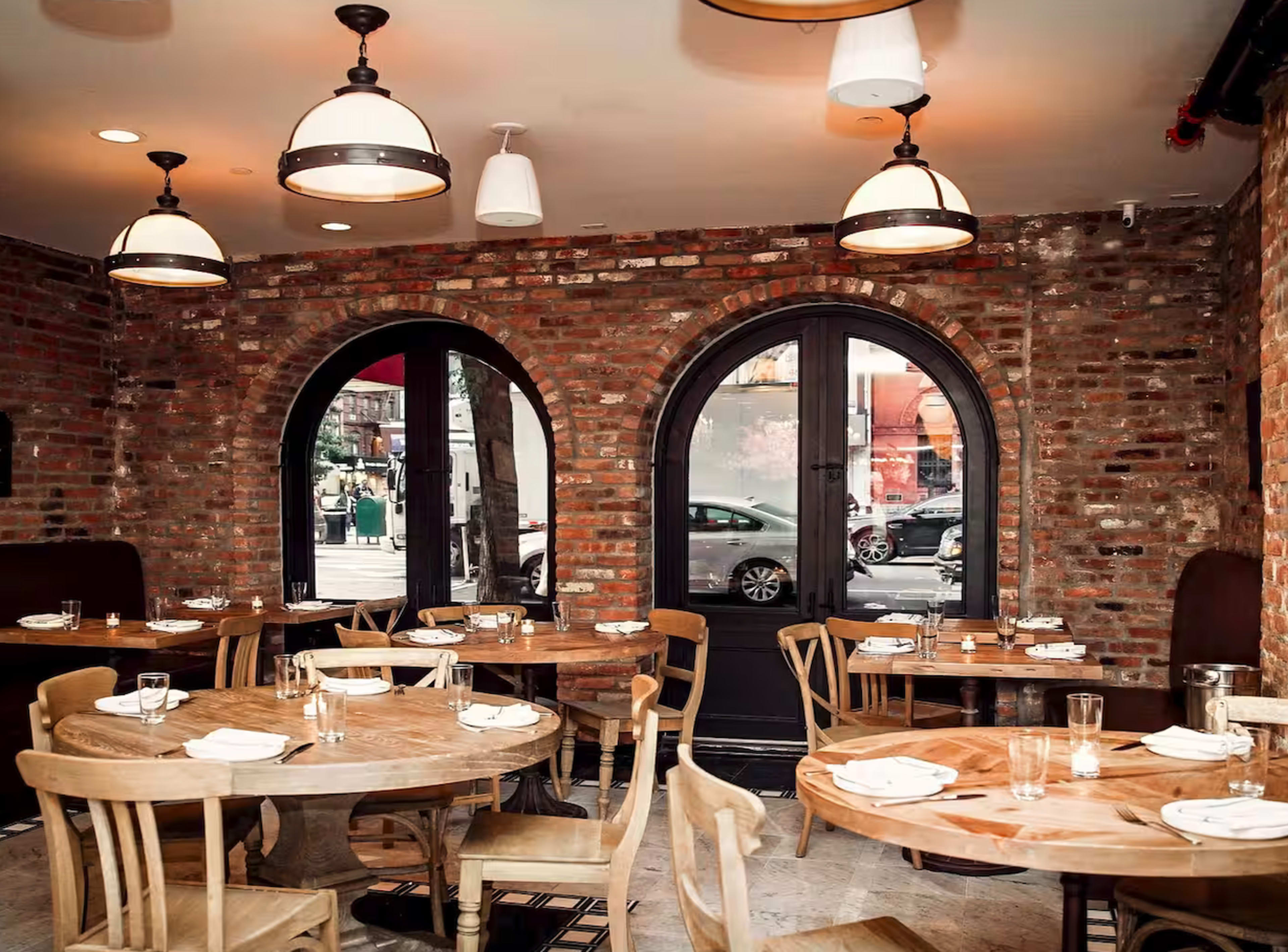 The image shows a rustic restaurant interior featuring round wooden tables and arched windows with a brick wall backdrop.