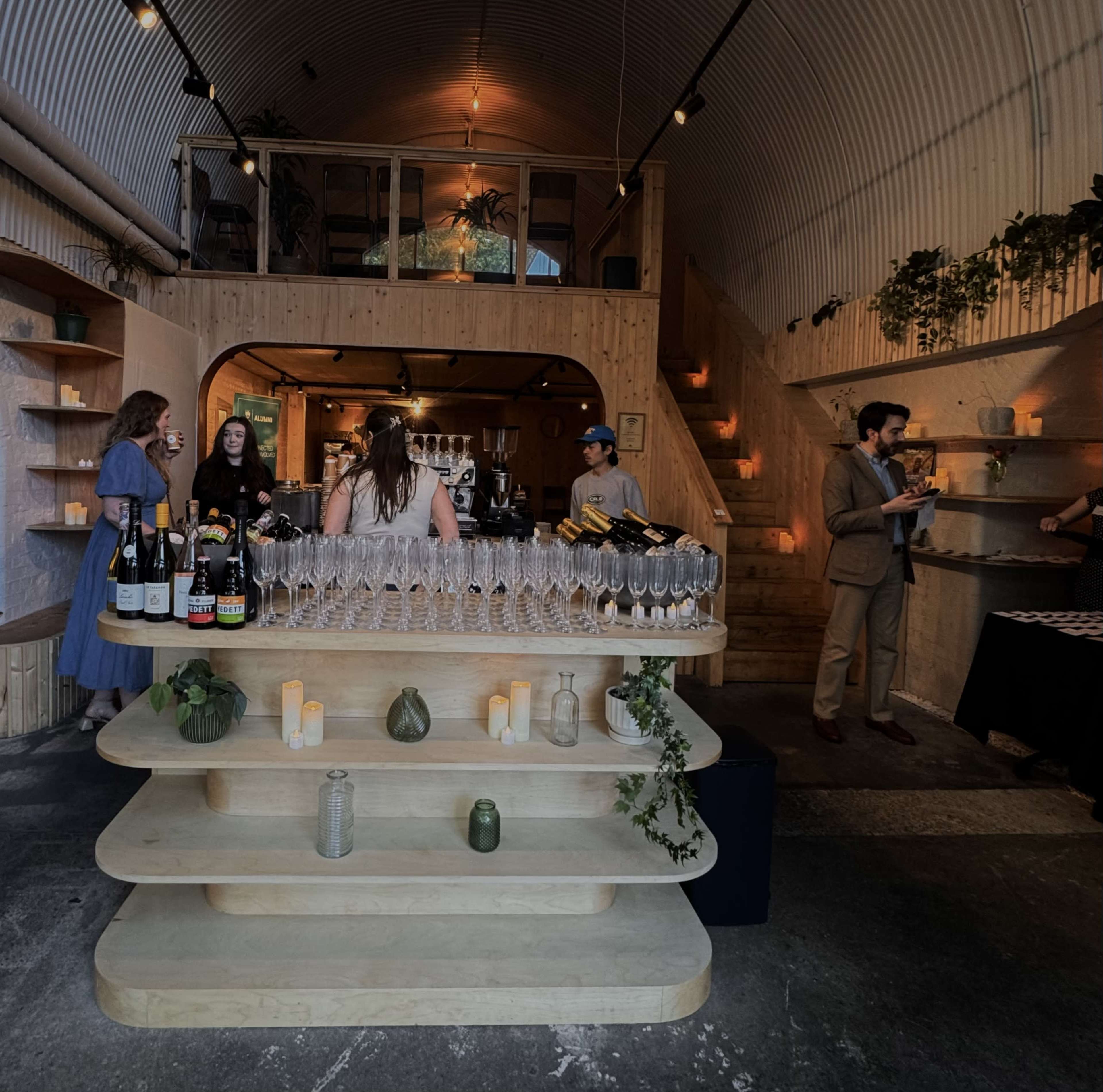 The image shows a modern bar interior with an organized display of wine bottles and champagne flutes, while patrons socialize and staff serve drinks in a spacious, well-lit space.