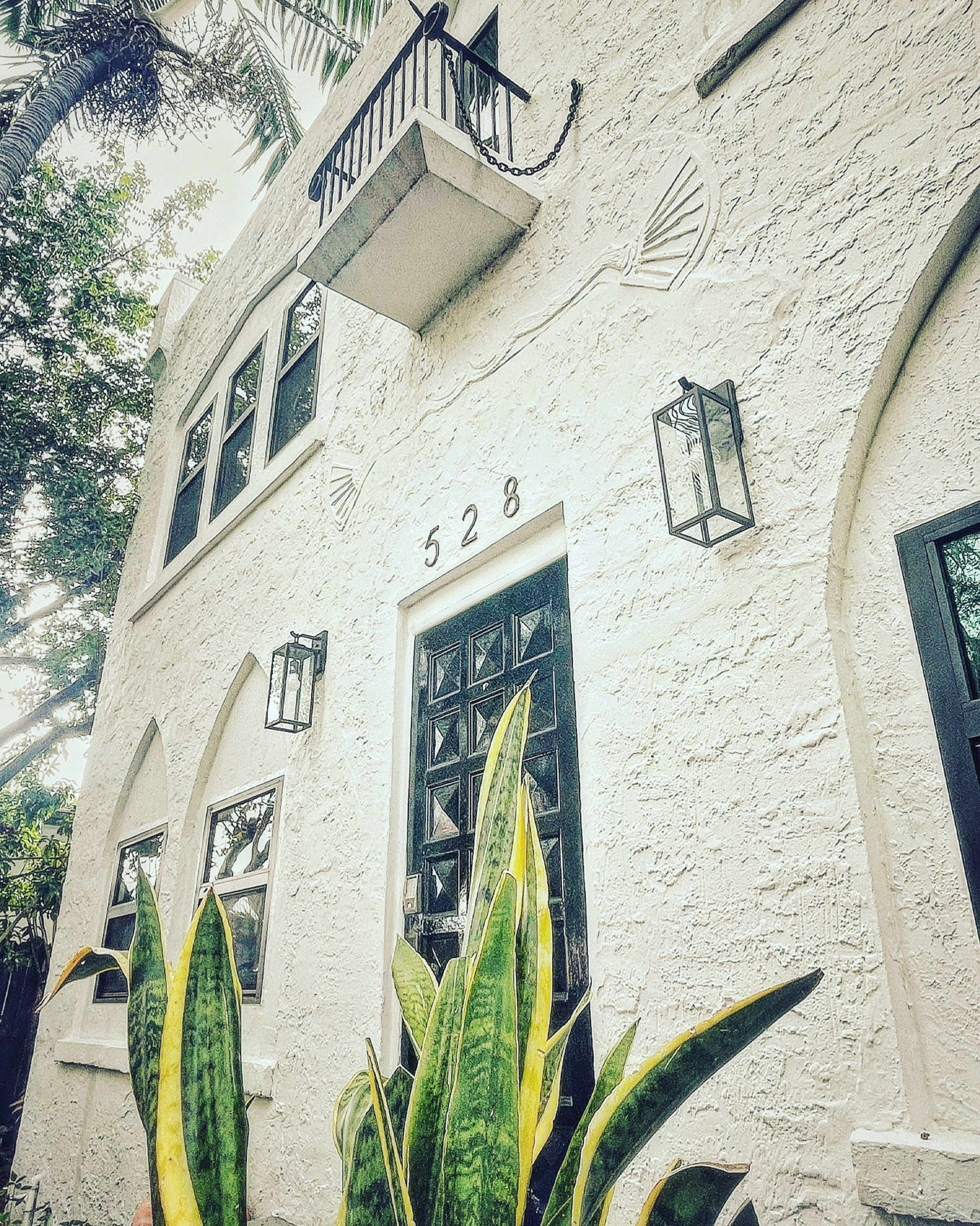 The image shows a white stucco building with arched windows, black doors, and green plants at the base.