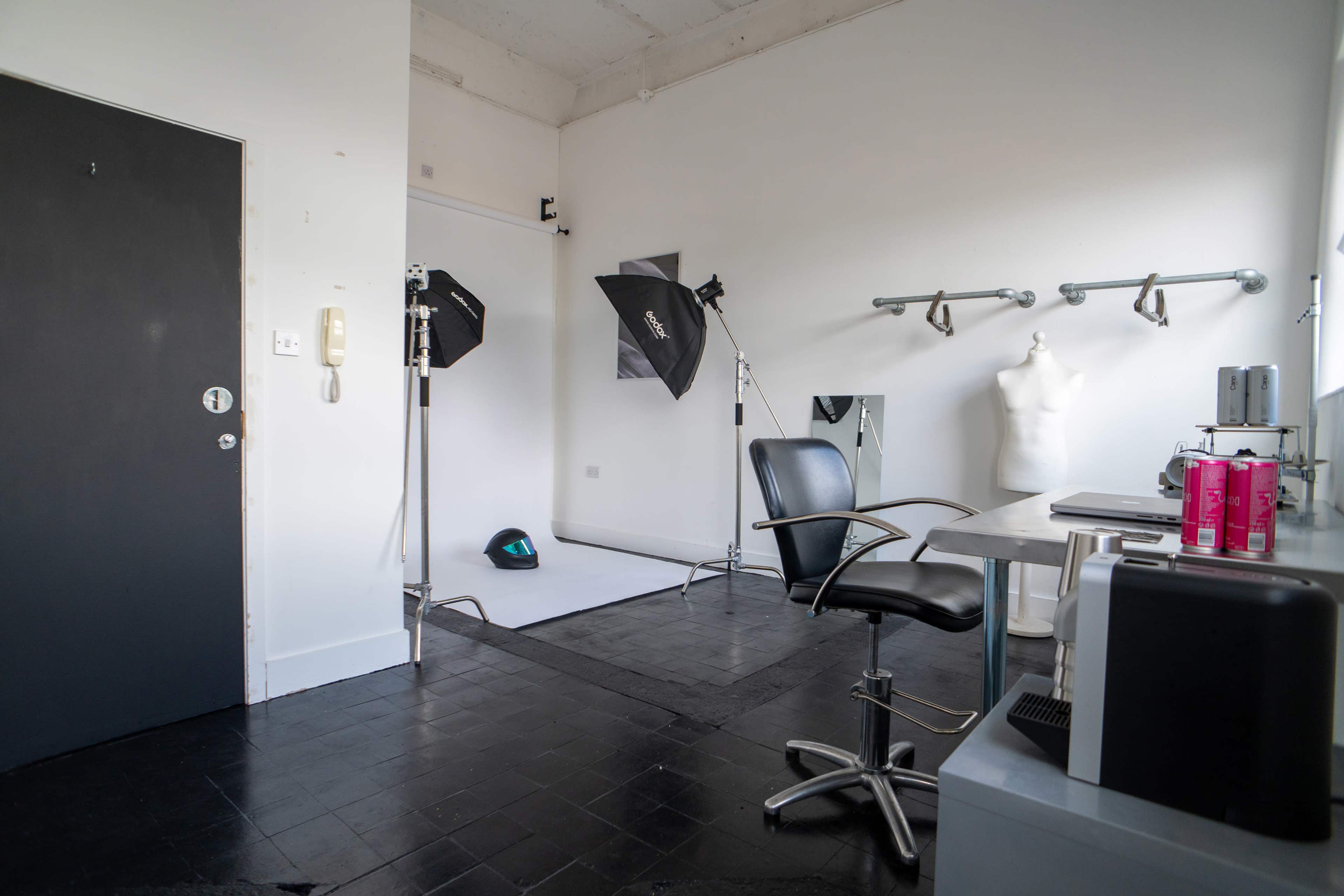 The image shows a photography studio featuring a black chair, a table with equipment, a backdrop setup, lights, and a mannequin.