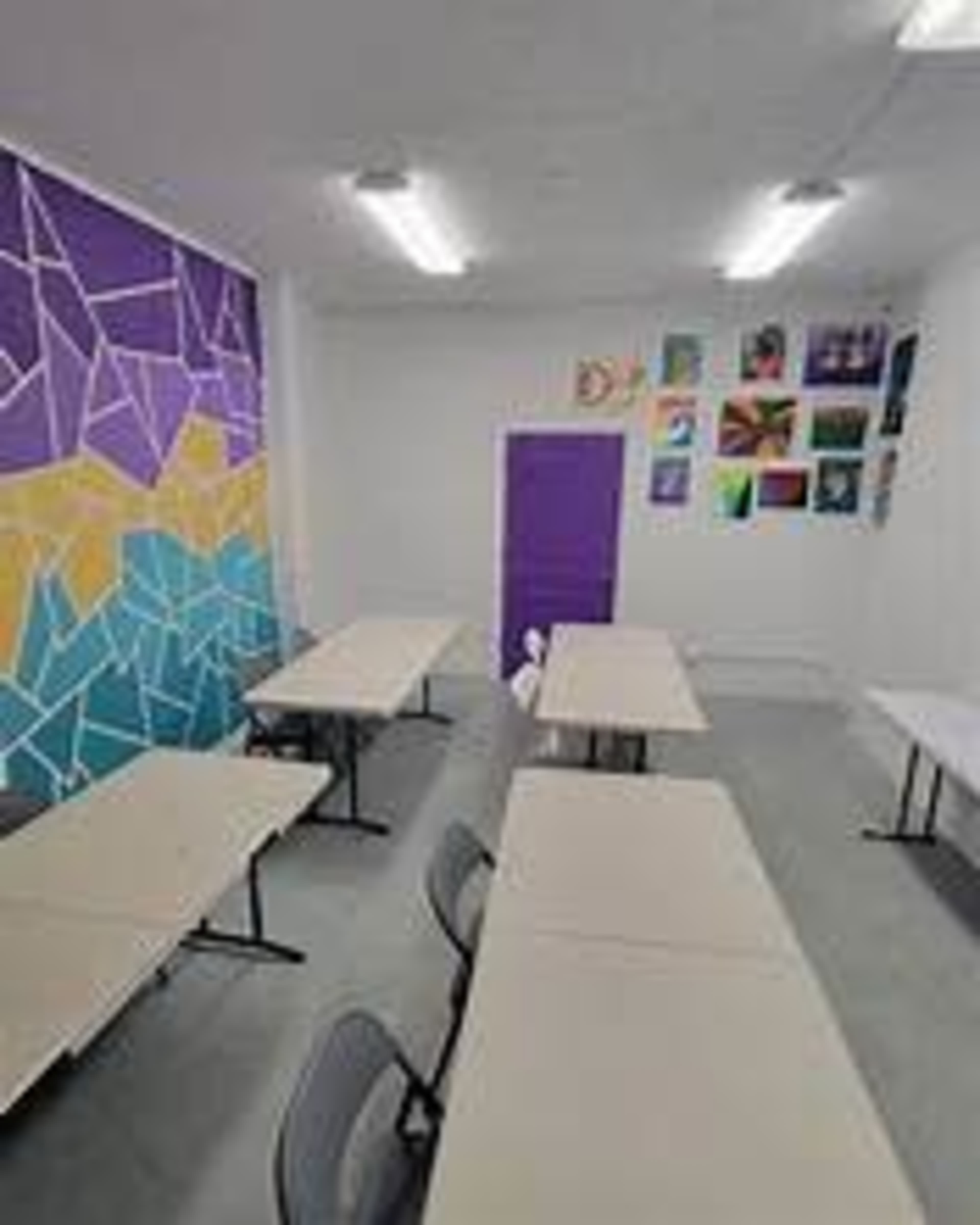 The image shows a classroom with several tables and chairs arranged neatly, a colorful mural on one wall, and artwork displayed on another.