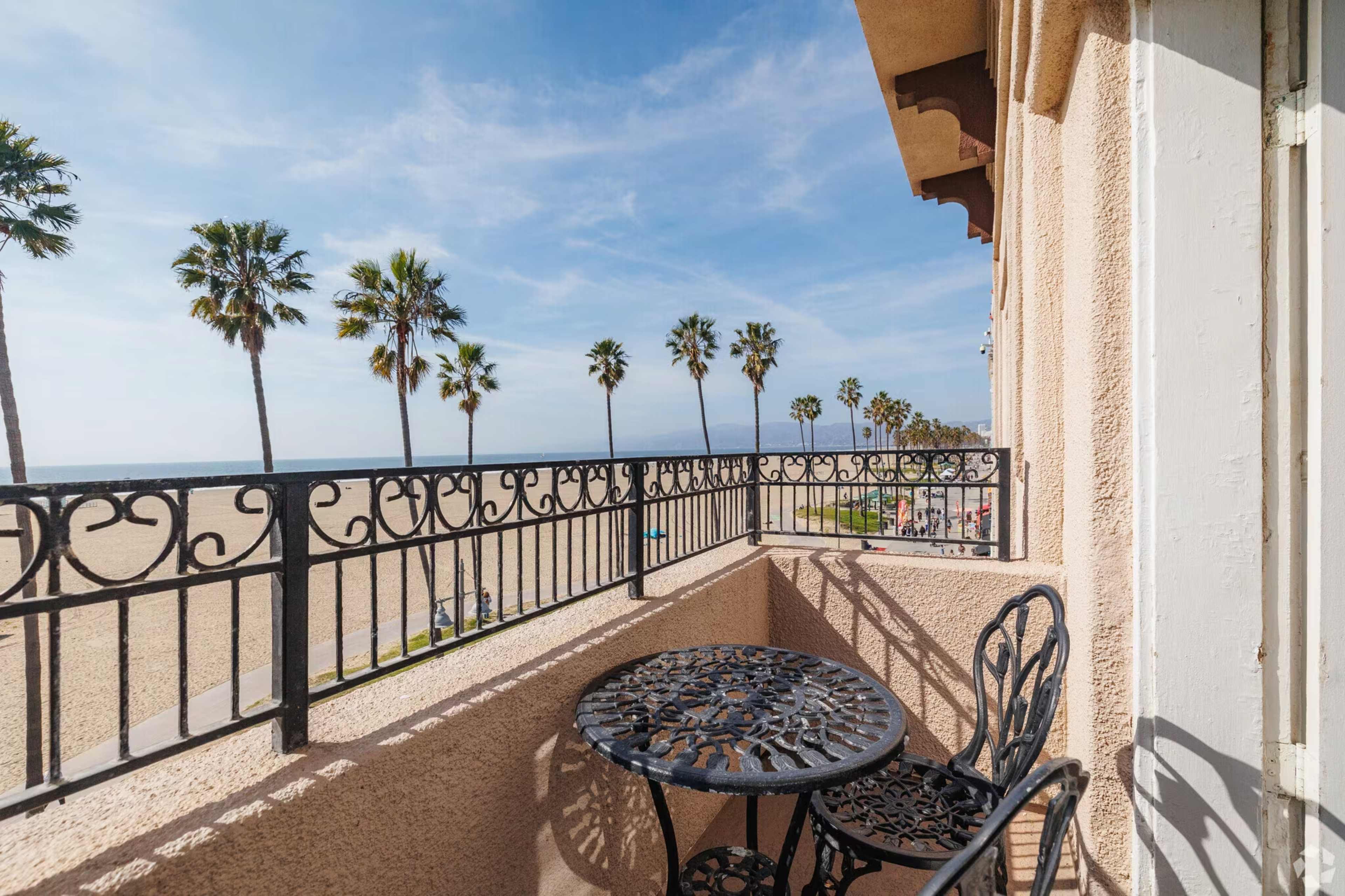 A balcony with a small table and chairs overlooks a sandy beach lined with palm trees and the ocean in the distance.