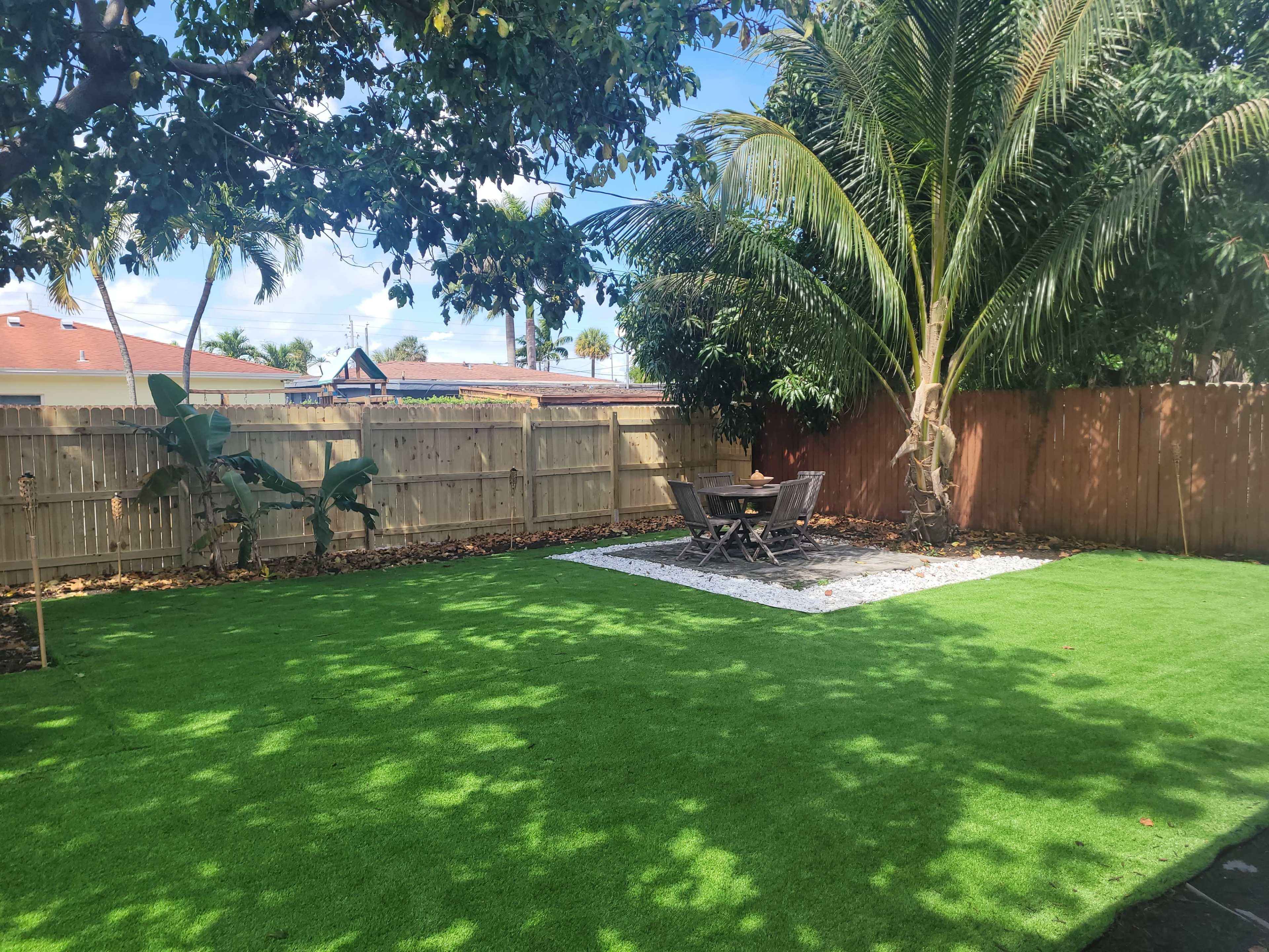 The image shows a grassy backyard with a stone patio surrounded by tropical trees and a wooden fence.