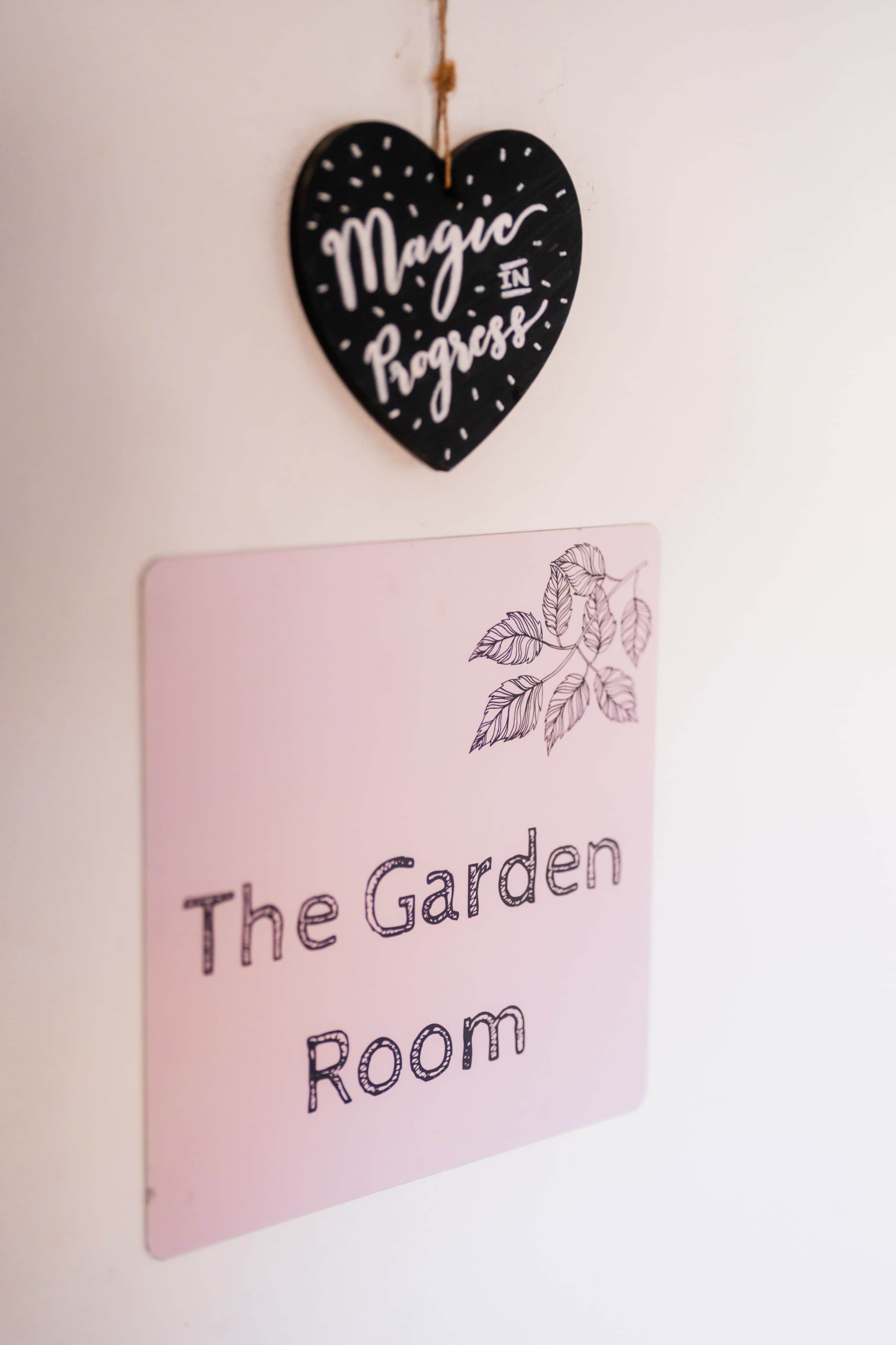 A heart-shaped sign reading "Magic in Progress" hangs above a square sign that says "The Garden Room" with decorative leaves.