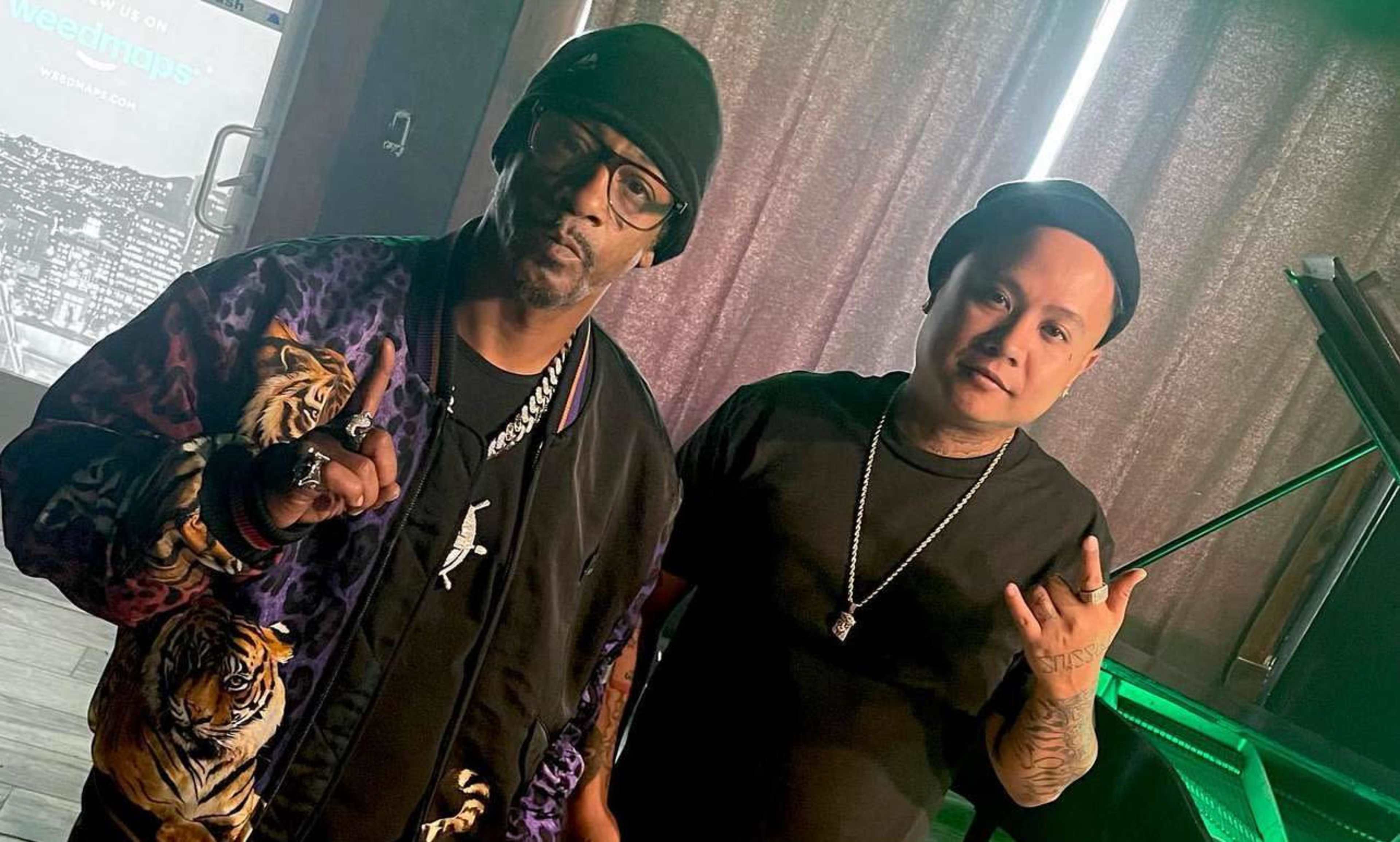 Two men pose for a photo indoors, one wearing a colorful jacket with tiger designs and the other dressed in a black shirt and beanie.