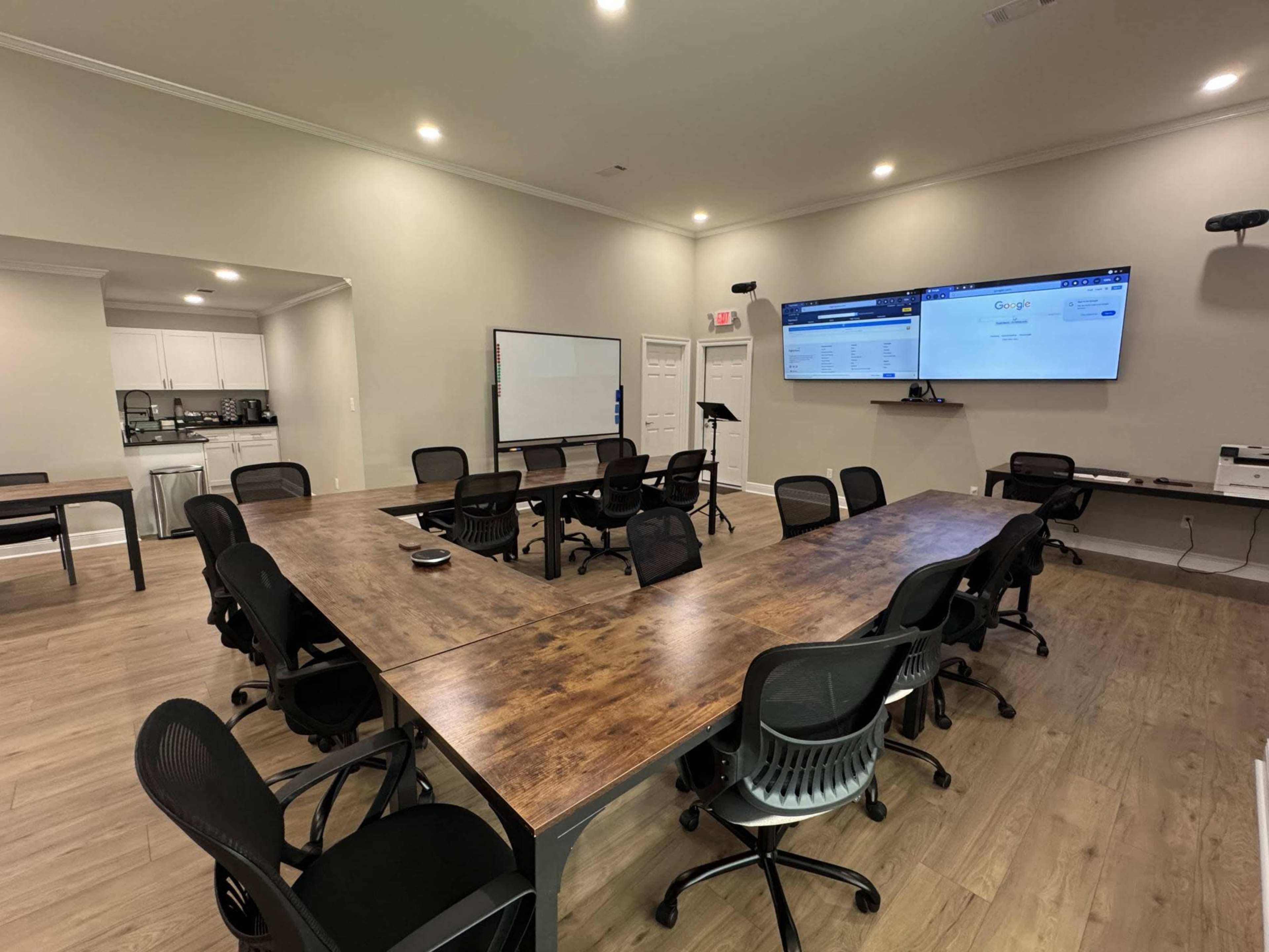 The image shows a modern conference room with a large wooden table arranged in a U-shape, surrounded by black office chairs, and equipped with a presentation screen on the wall.