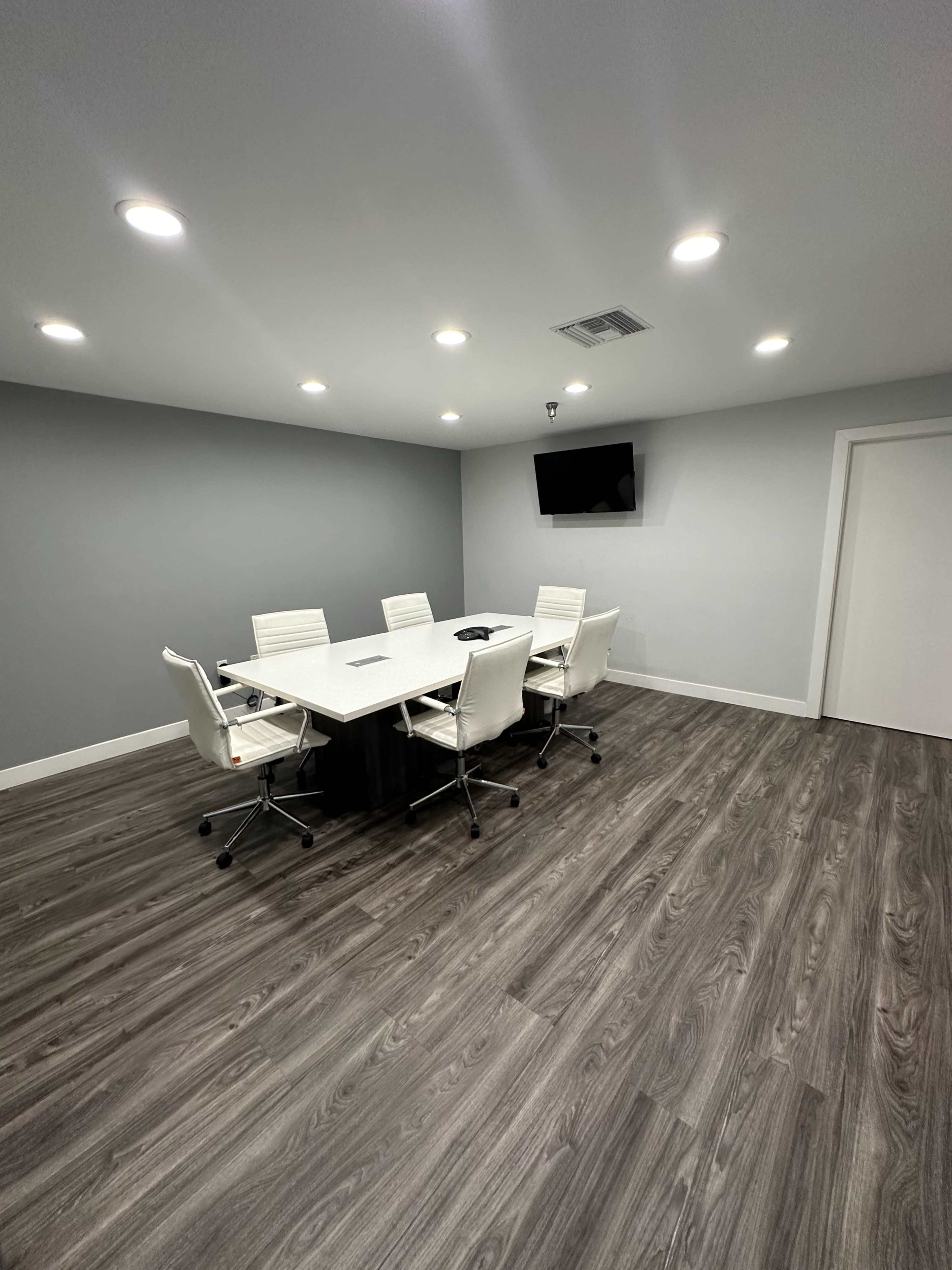 A modern, minimalist conference room features a rectangular table surrounded by white chairs, with a television mounted on the wall and recessed lighting above.