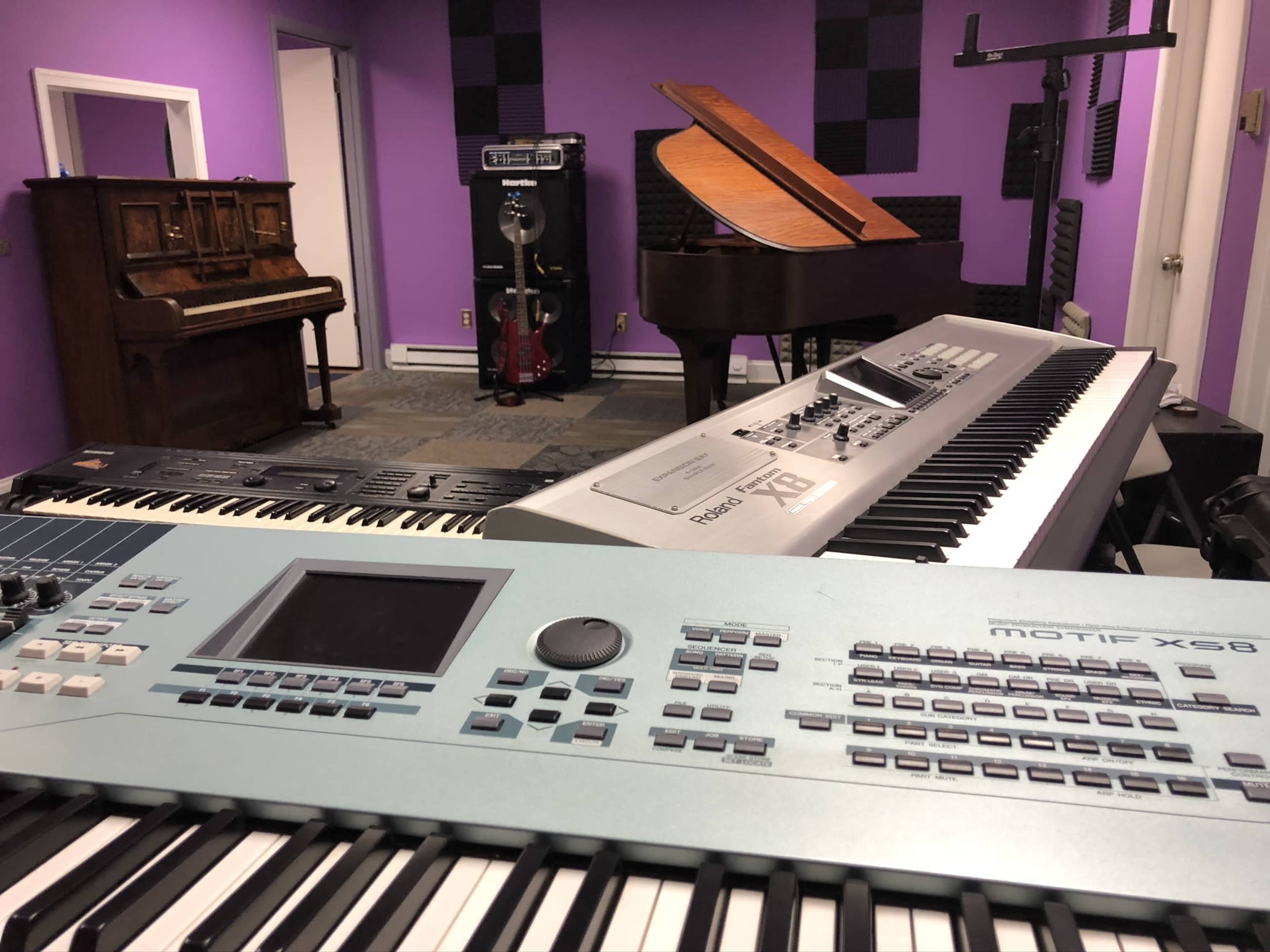 A music studio with various instruments, including a grand piano, an upright piano, and multiple keyboards, set against purple walls.