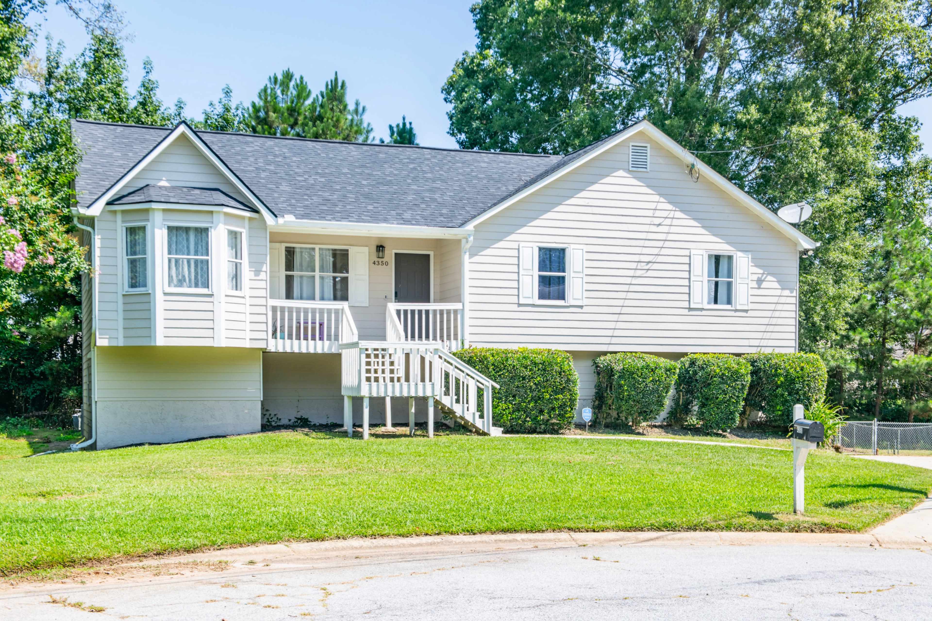 A single-story house with a light gray exterior and a front porch is situated on a green lawn.