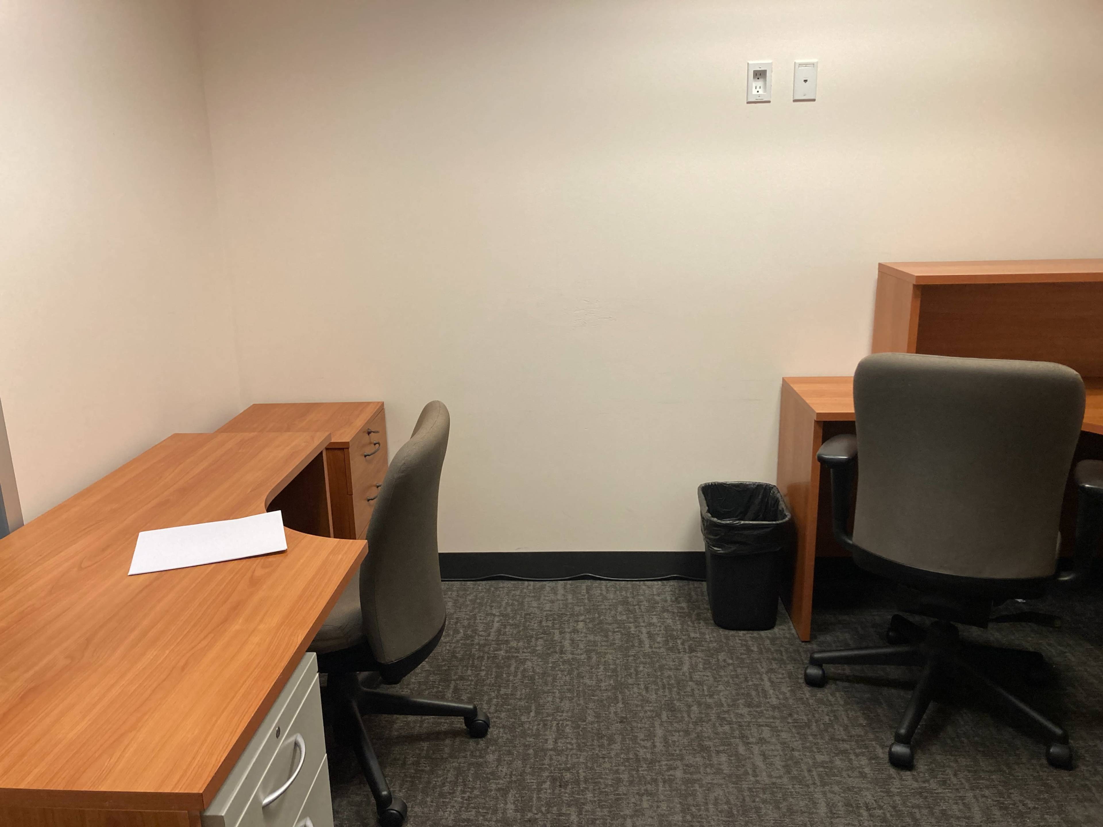The image shows a sparse office space with two wooden desks, two office chairs, and a trash can.