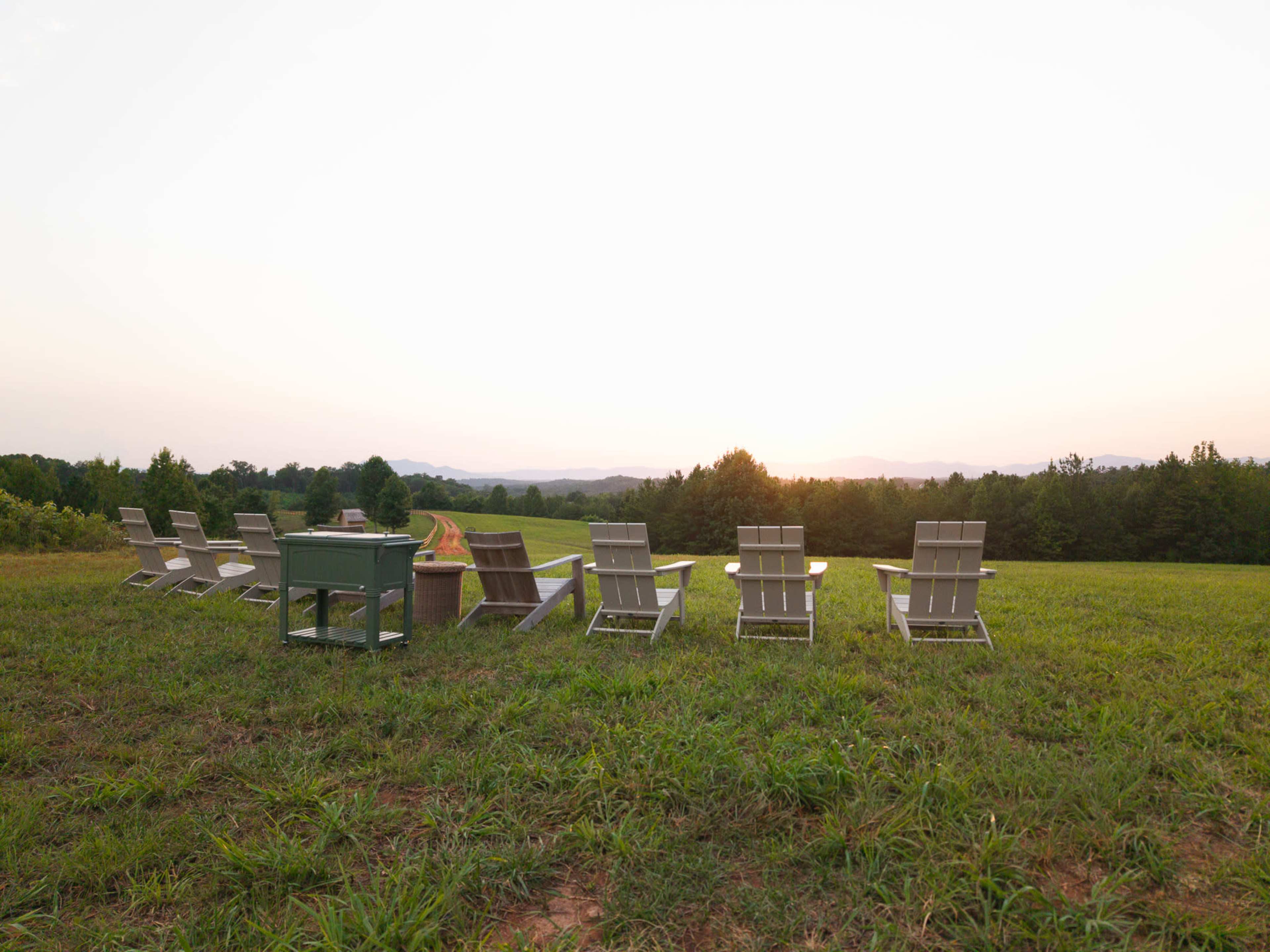 A row of white lawn chairs faces a sunset over a grassy field and hills.