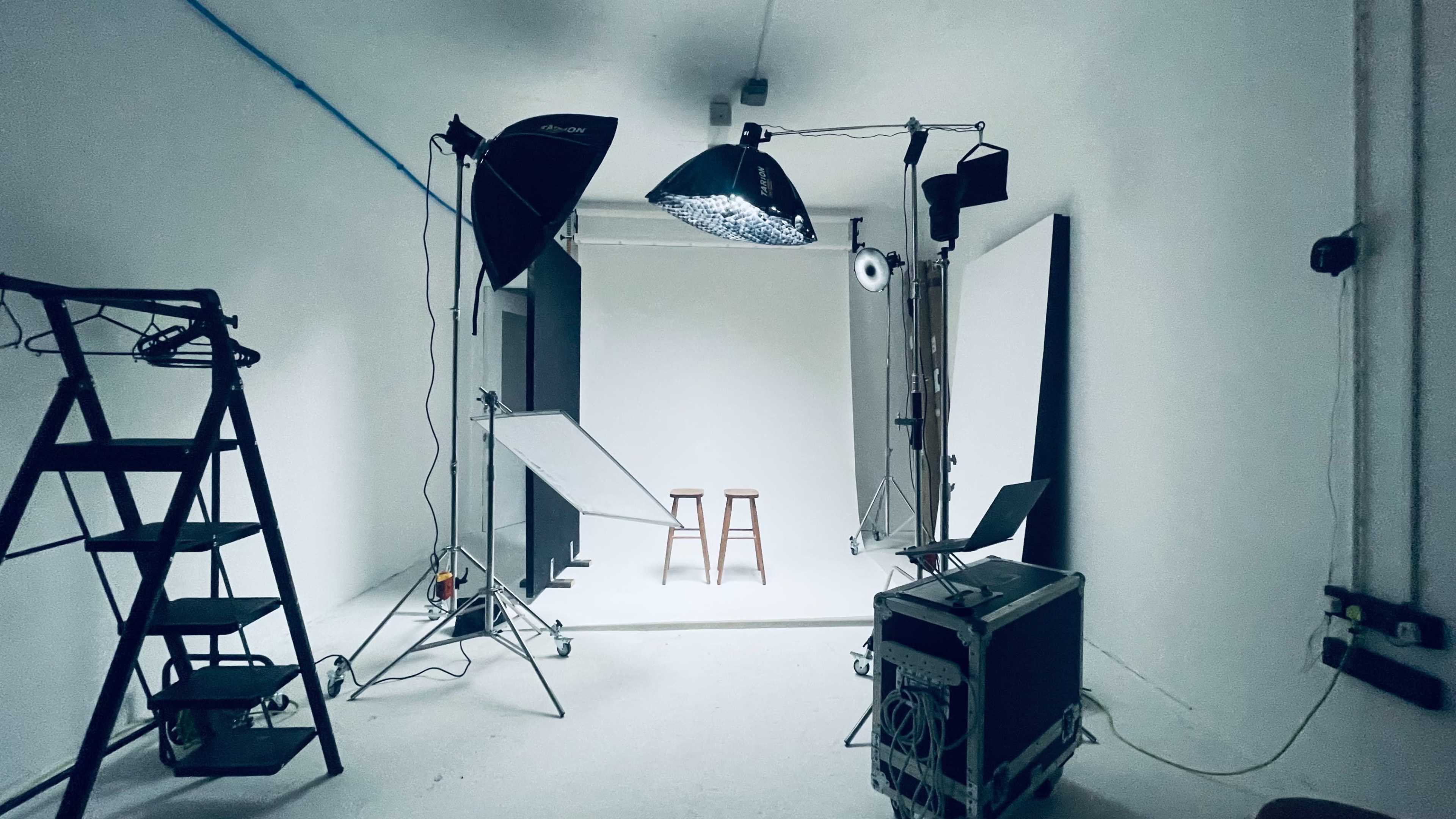 The image shows a photography studio setup with two wooden stools, various lighting equipment, and backdrops positioned on the floor.