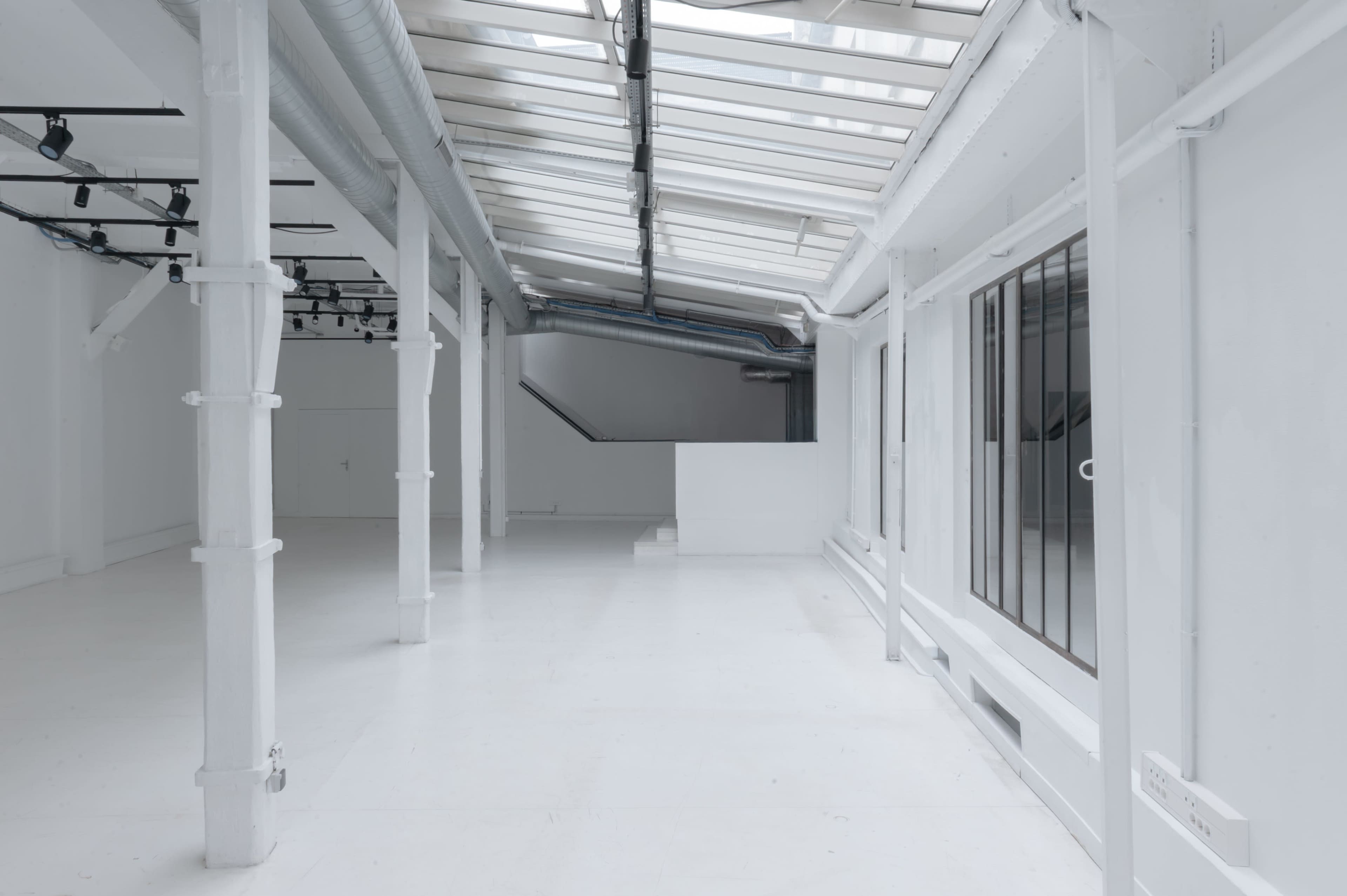 The image shows a spacious, bright room with white walls and a glossy floor, featuring large windows and a minimalist design.