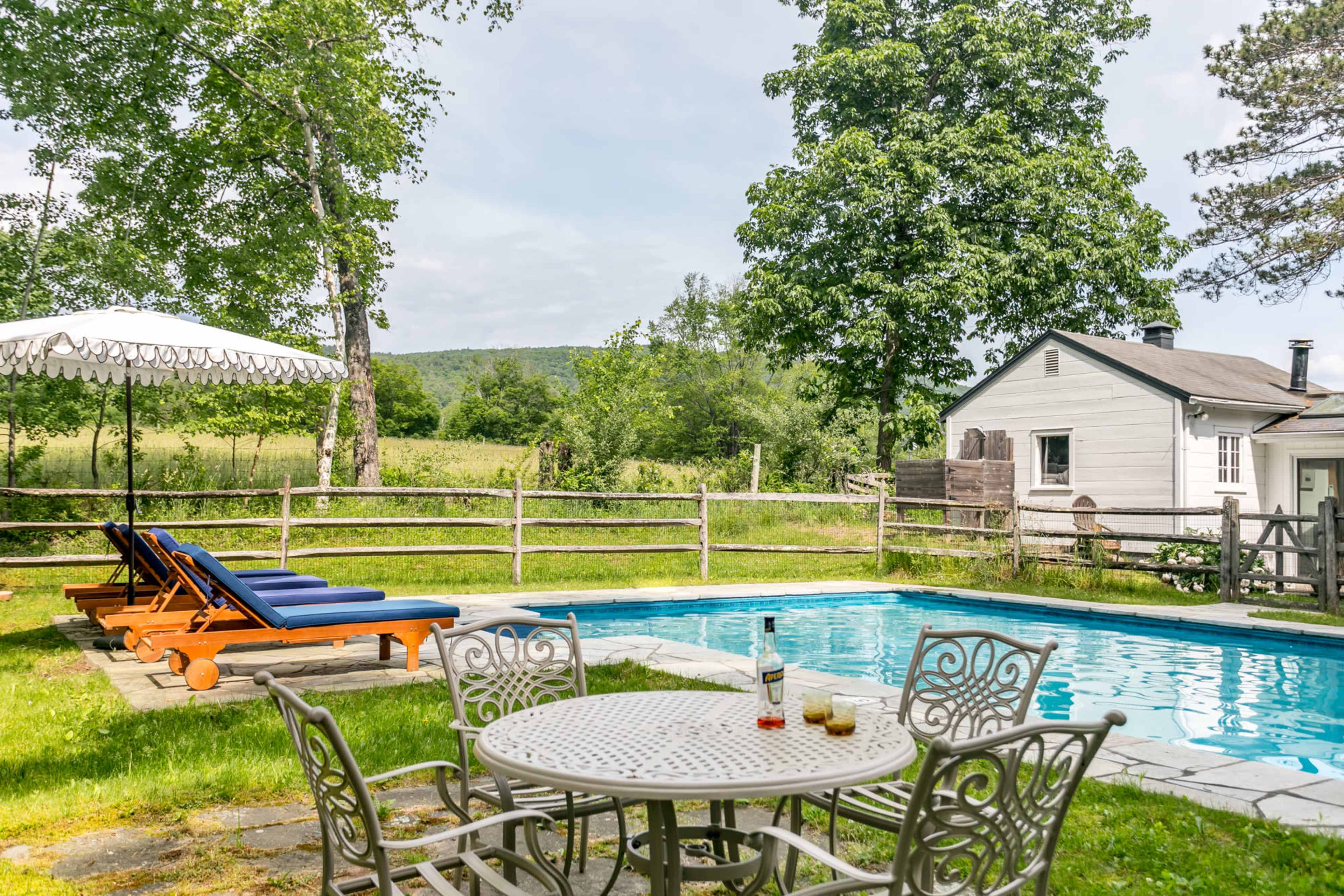 The image depicts a serene backyard with a swimming pool surrounded by lounge chairs, a table with drinks, and a farmhouse set against a scenic landscape.