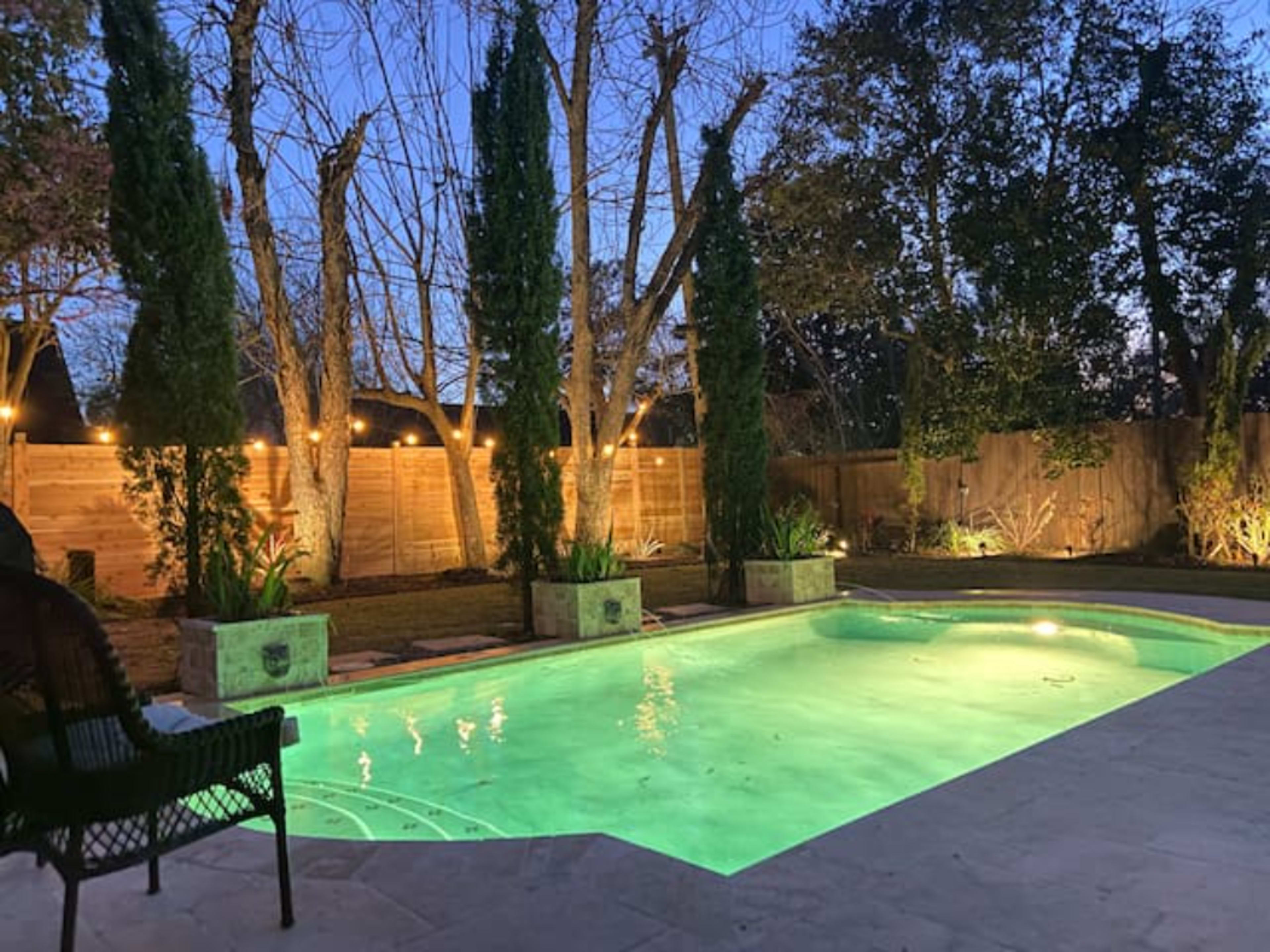 A well-lit backyard features a swimming pool surrounded by greenery and trees at dusk.