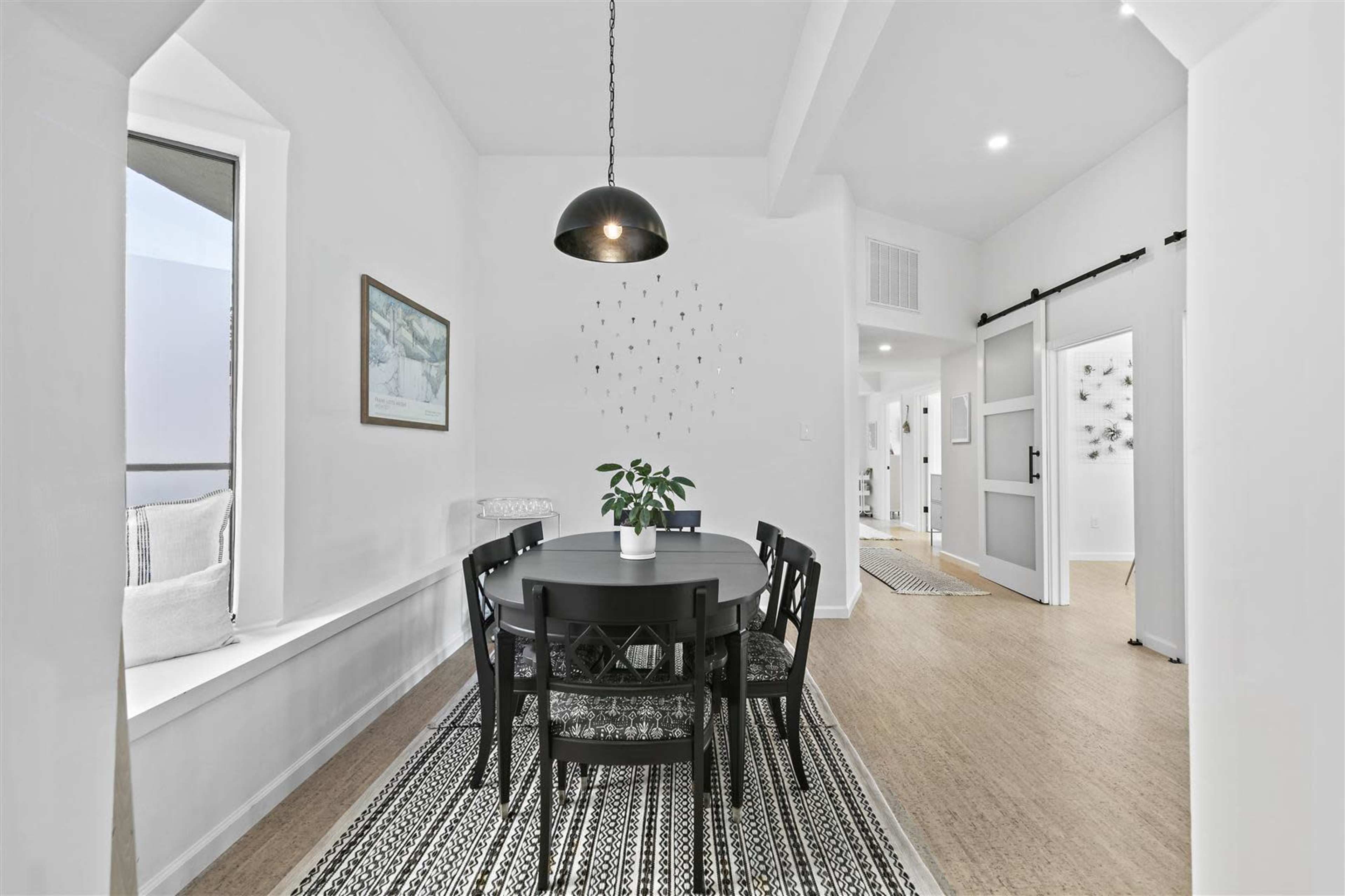 A dining area features a round black table surrounded by six chairs, with large windows and a potted plant on the table.