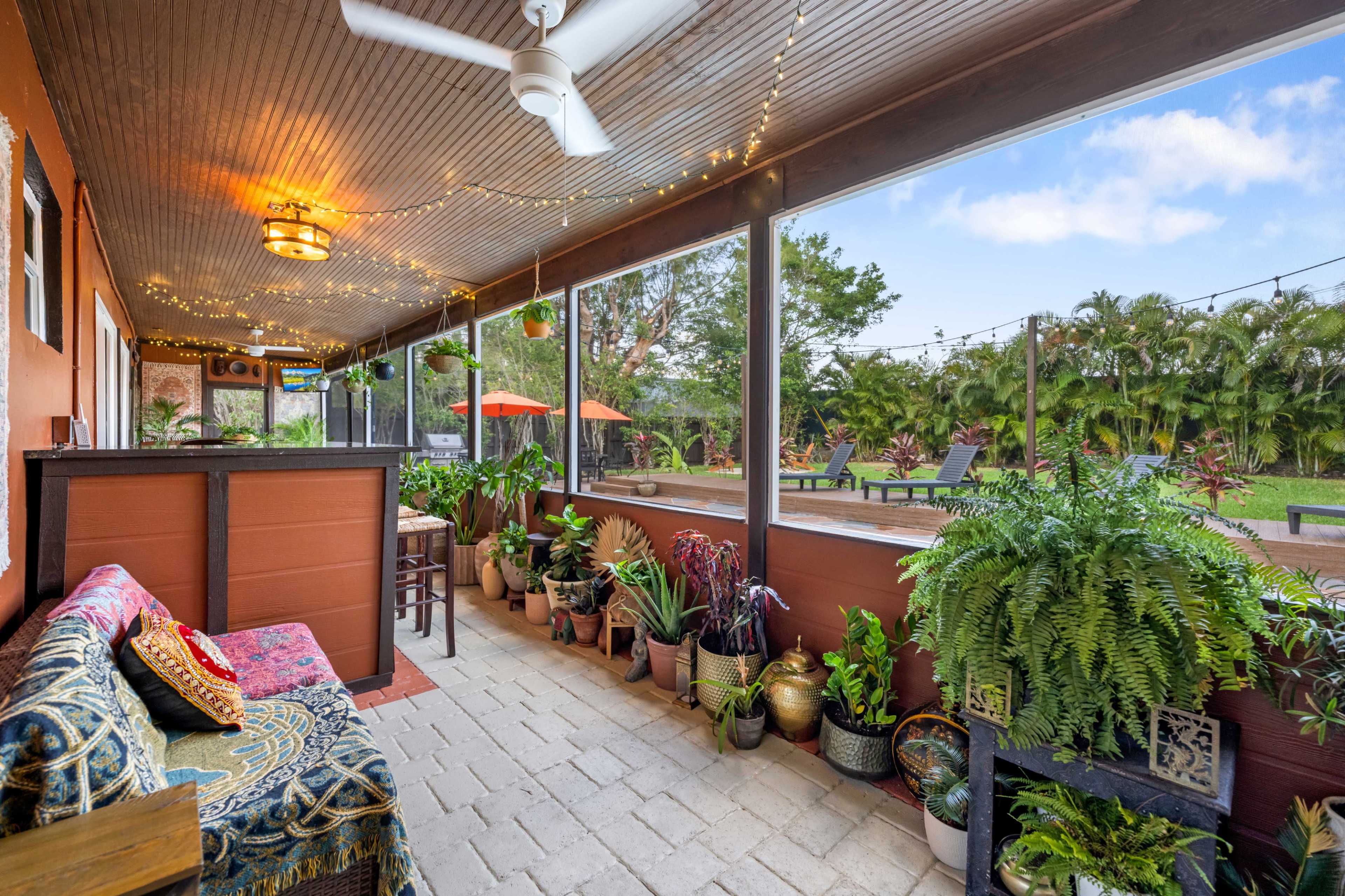 A covered porch adorned with string lights, potted plants, and a seating area, overlooking a landscaped yard.