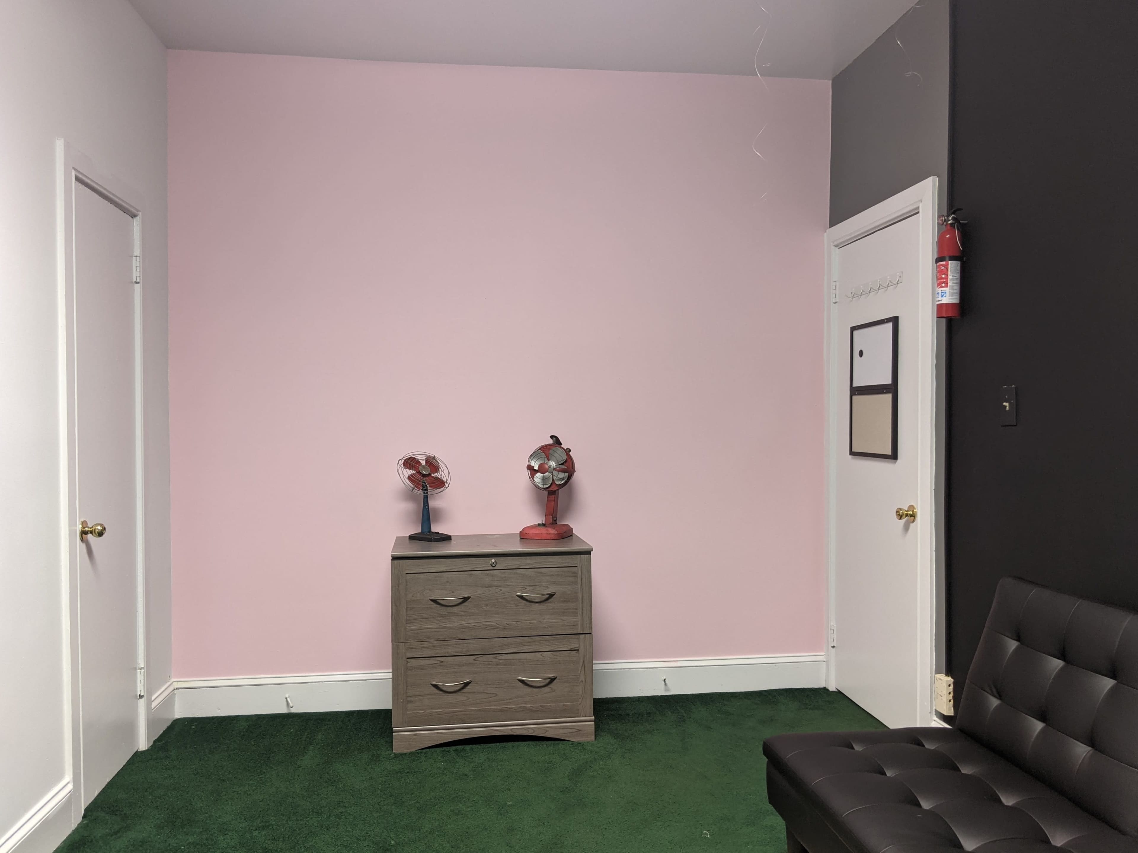The room features a pink wall, a gray dresser with two small fans on top, and a dark wall opposite a green carpet.