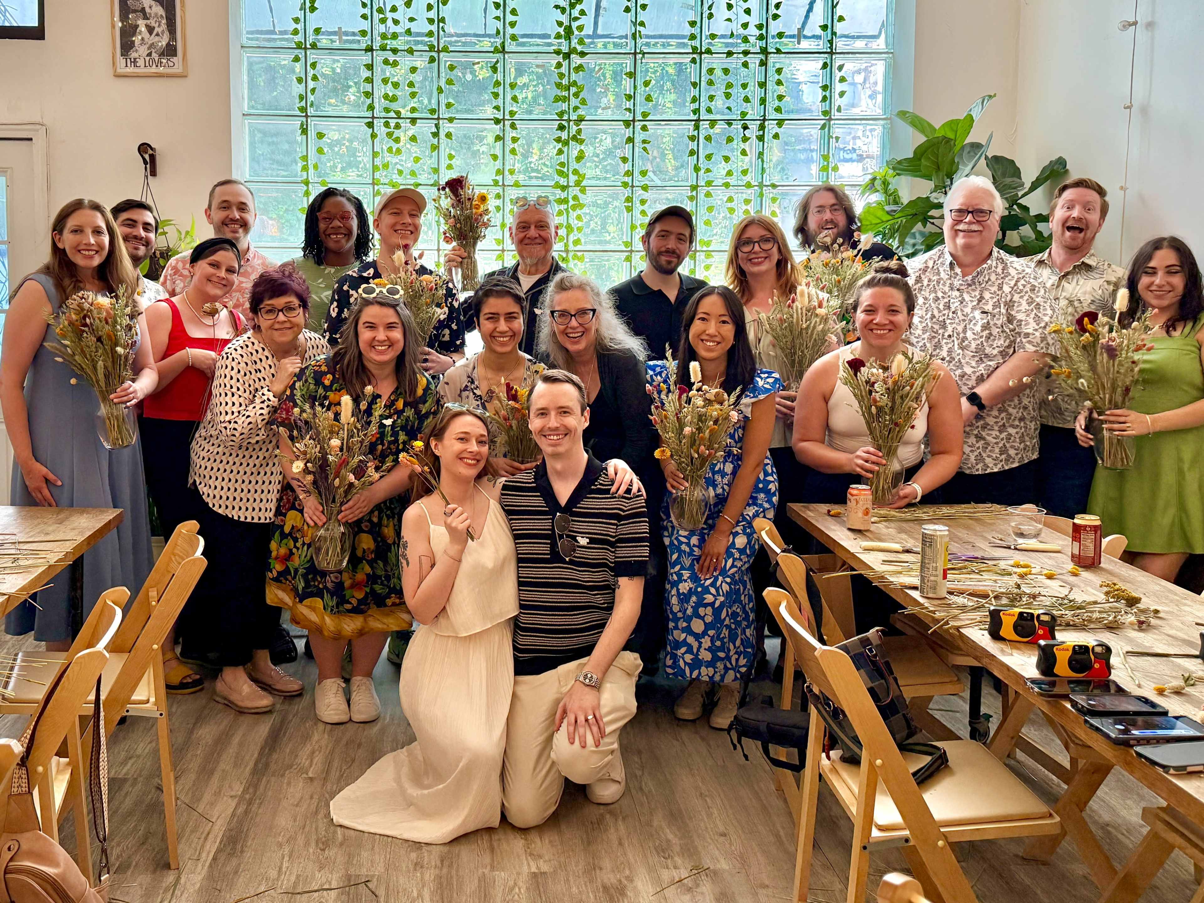 A diverse group of people poses together, holding bouquets of dried flowers, in a bright, plant-filled room with large windows.