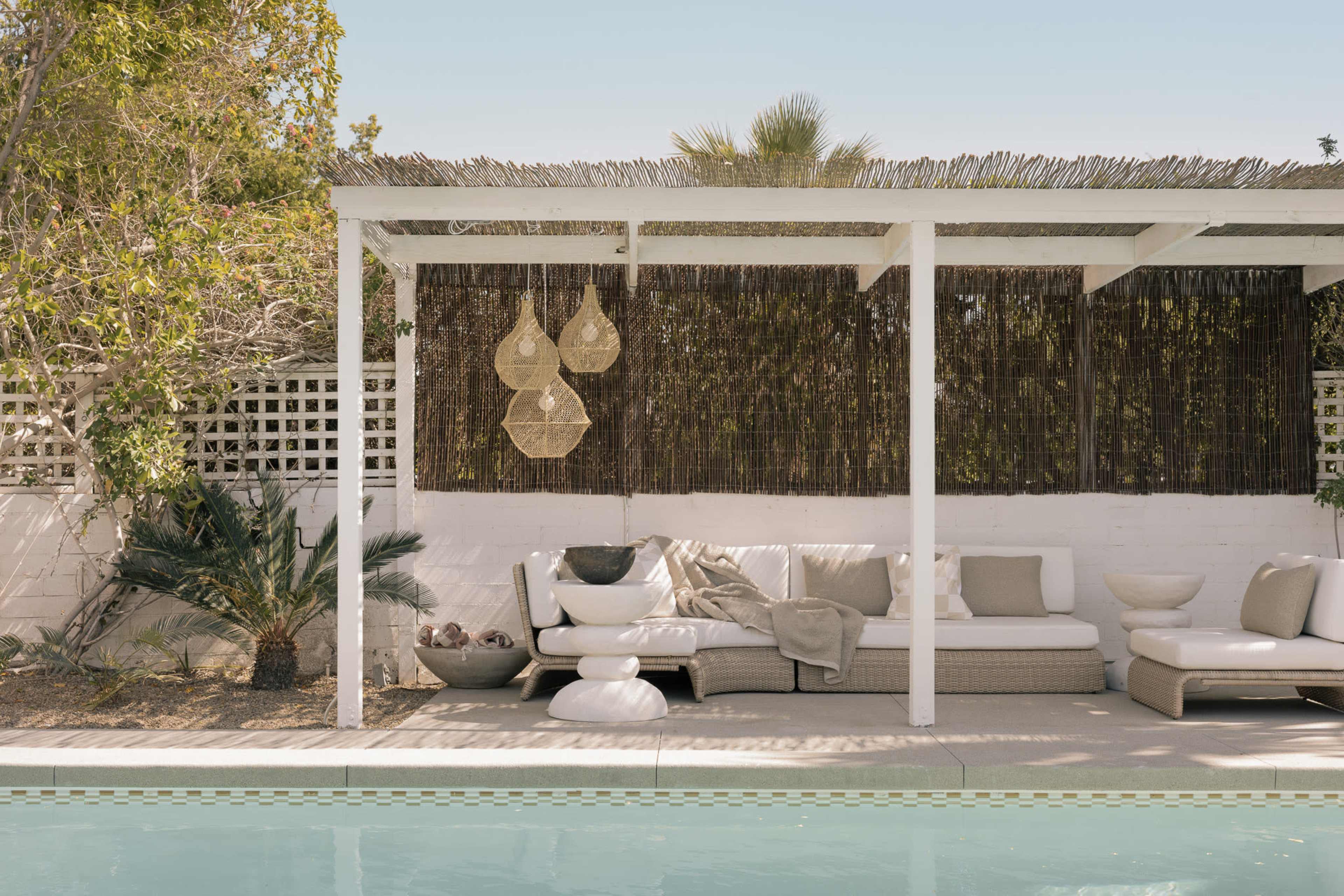 A white patio area with a sofa and decorative elements overlooks a swimming pool, framed by tall plants and a bamboo screen.