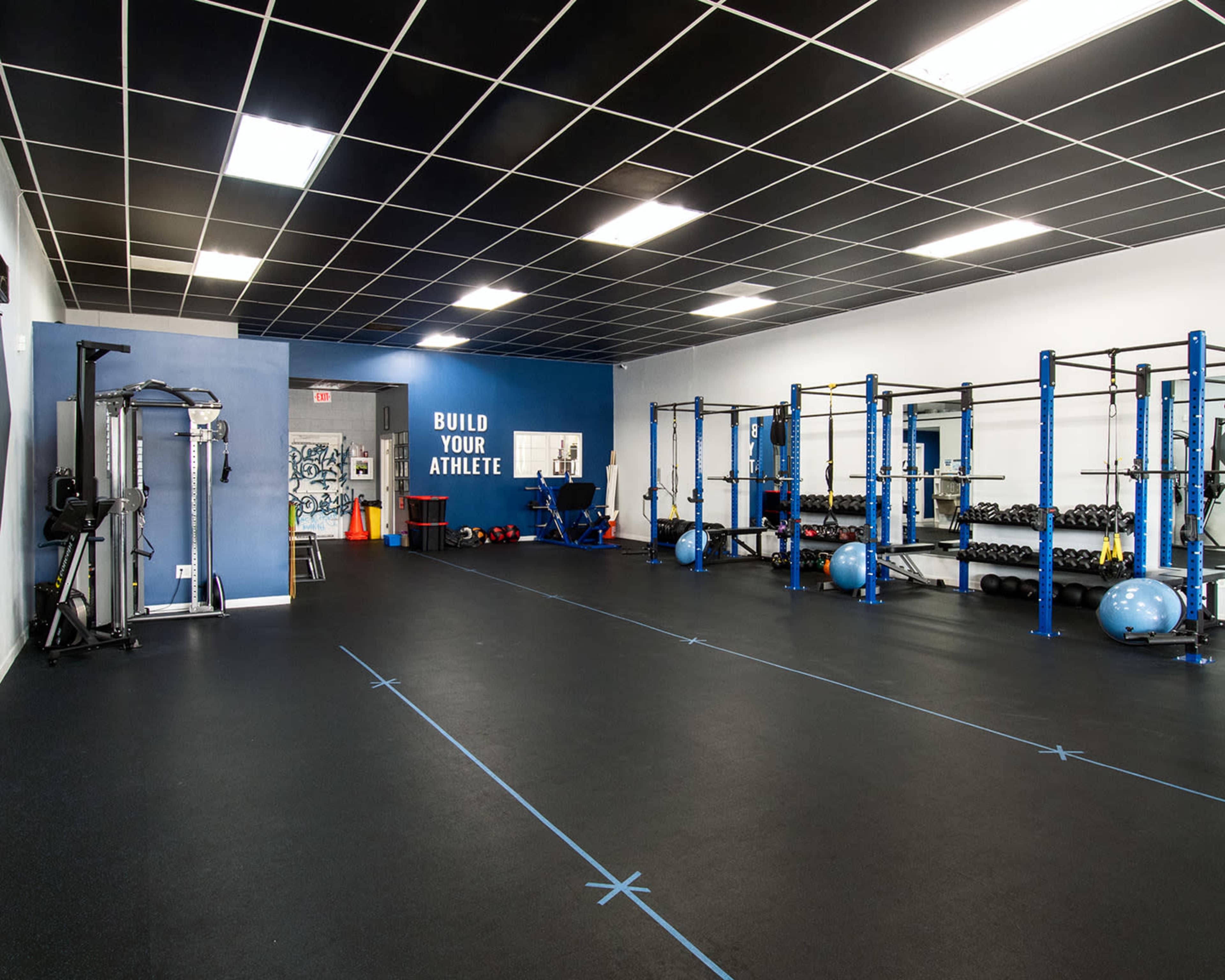 A spacious gym interior featuring weightlifting equipment, blue walls, and a large sign that reads "BUILD YOUR ATHLETE."