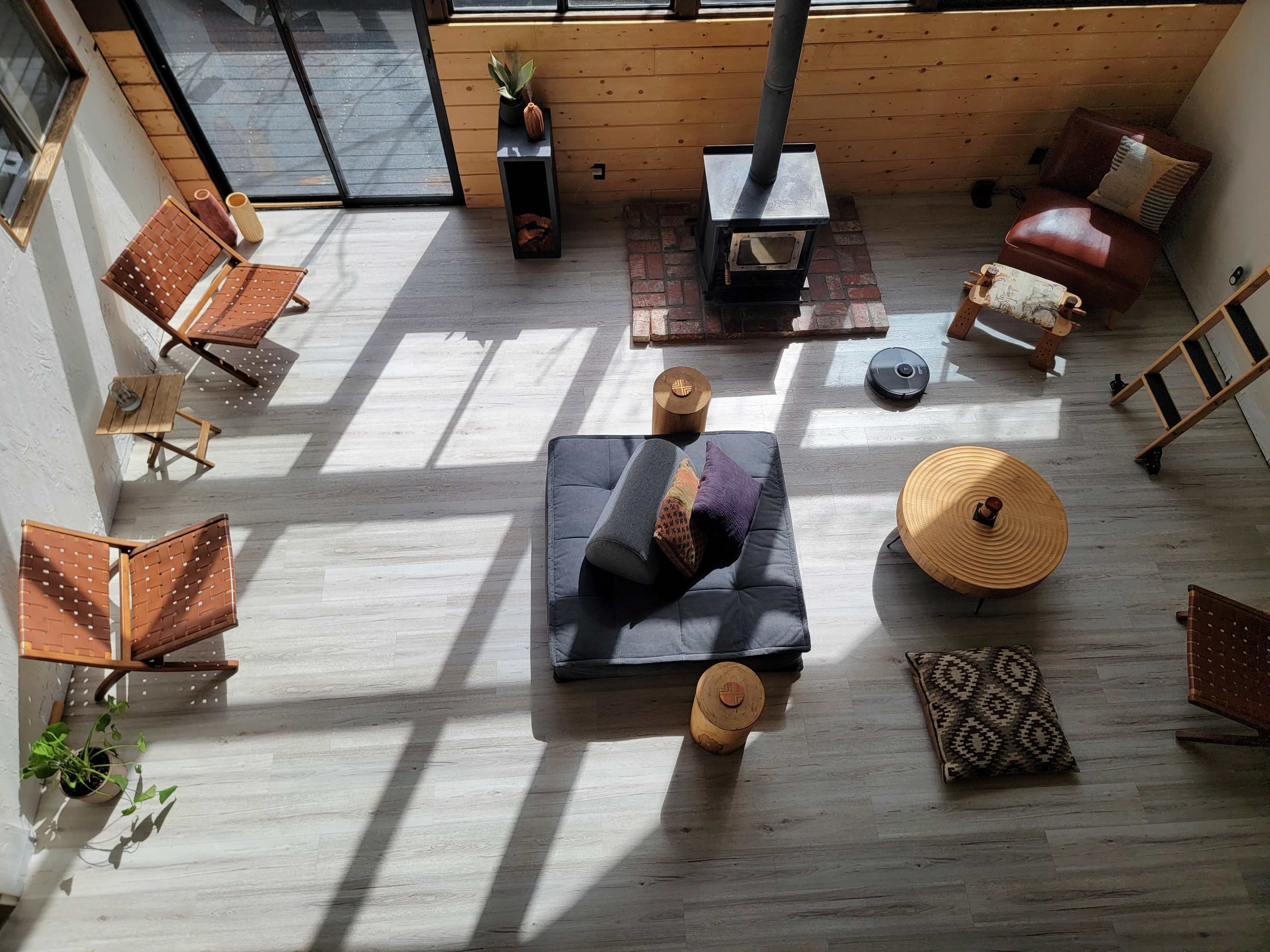 The image shows a spacious, well-lit living area with a wood-burning stove, a large black sofa, and various seating options, including wooden chairs and a round coffee table.