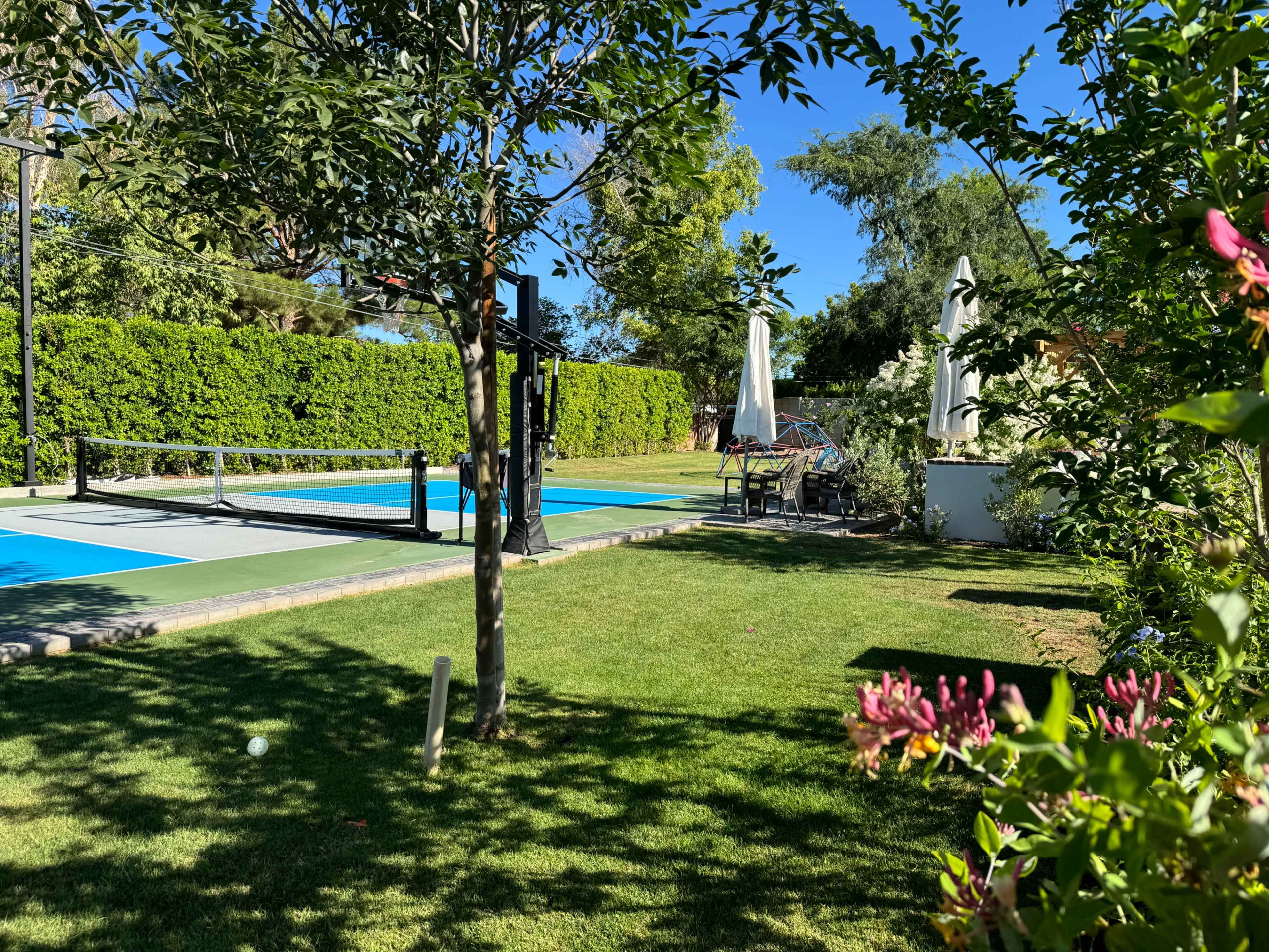 A landscaped garden with a tennis court on the left, a seating area under an umbrella on the right, and greenery surrounding the area.