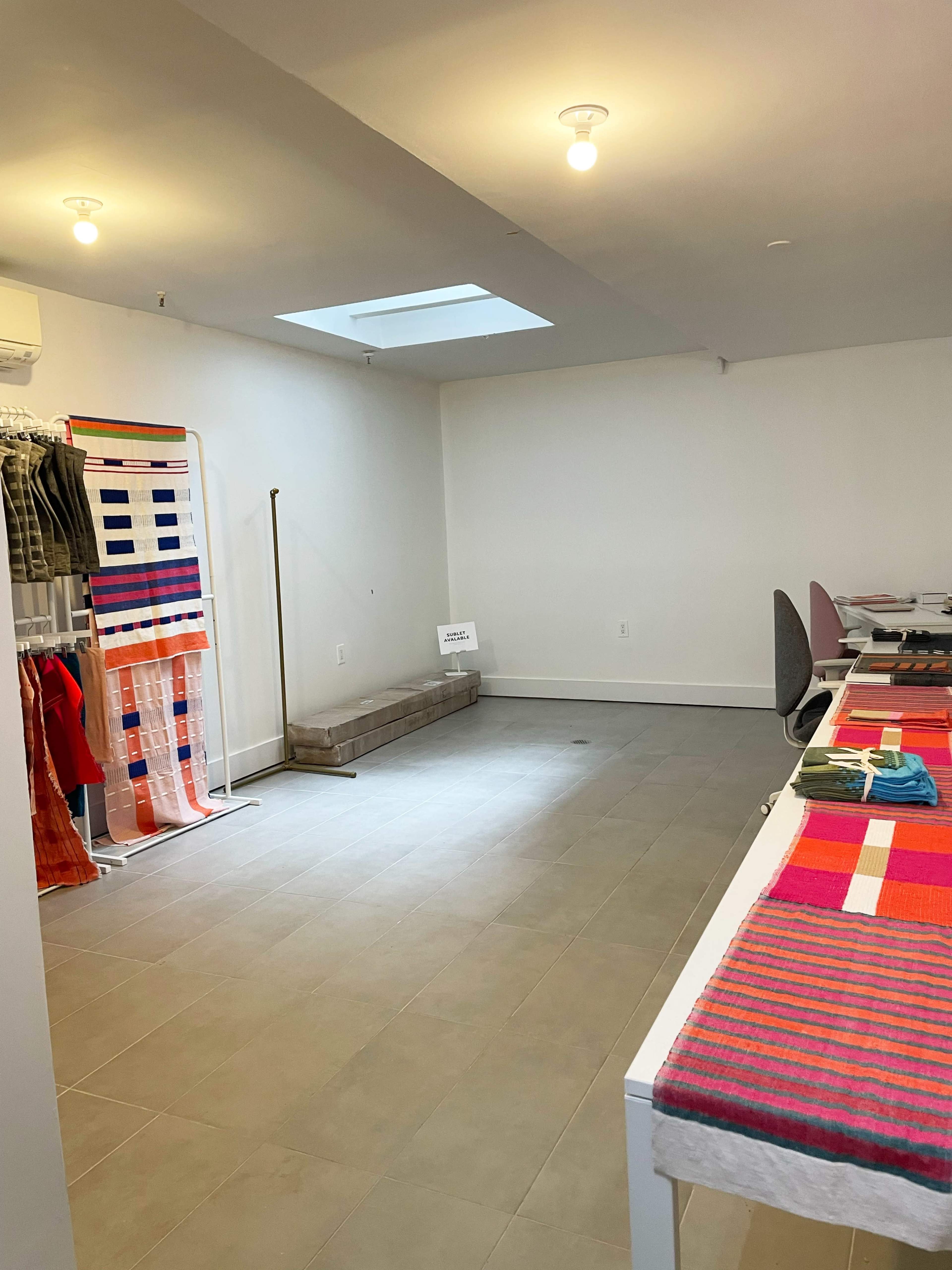 The image shows a spacious, minimally decorated boutique with colorful textiles displayed on tables and hanging on a rack, illuminated by overhead lights.