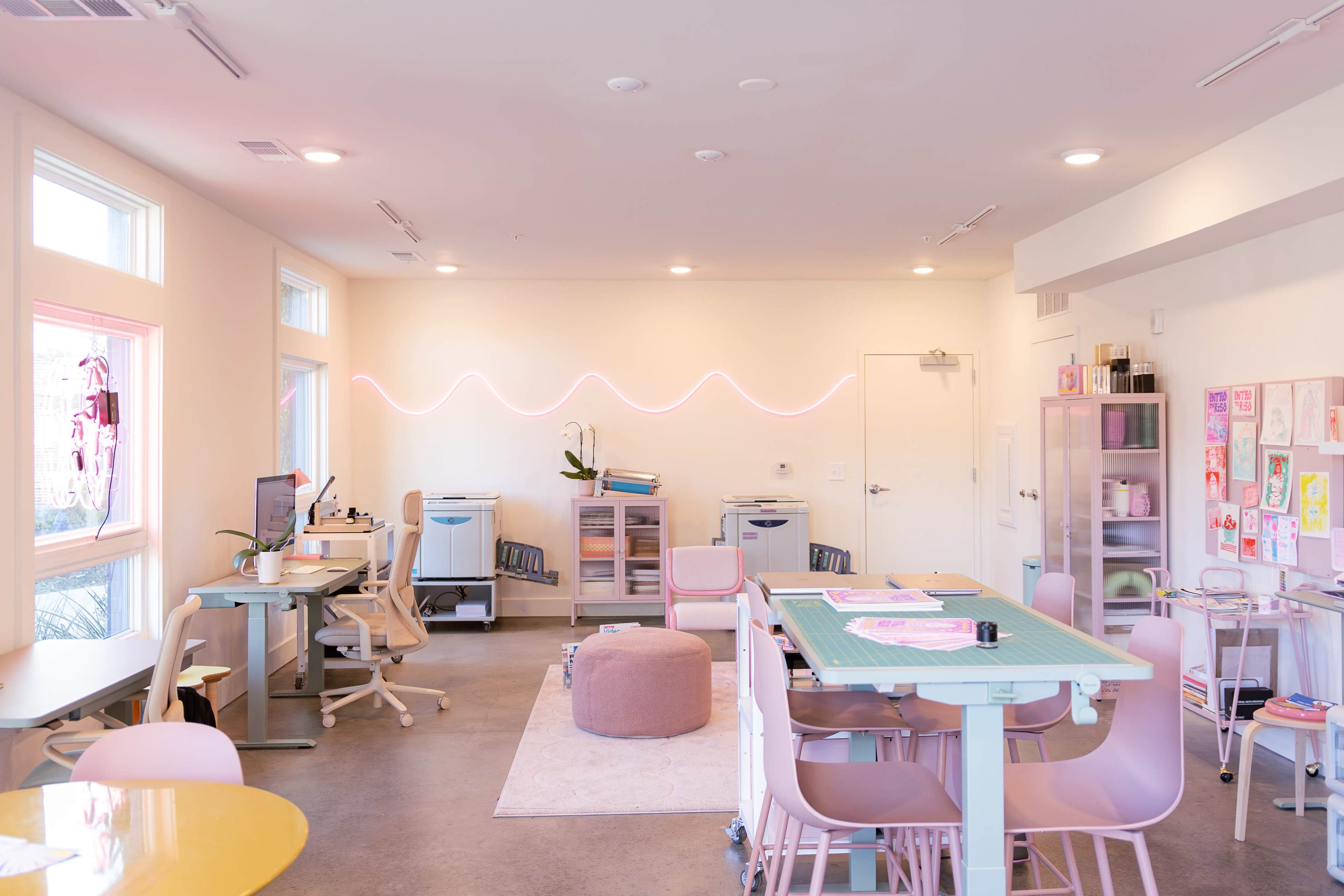 The image shows a bright, modern workspace with light-colored walls, a green cutting table, and various workstations equipped with computers and art supplies.