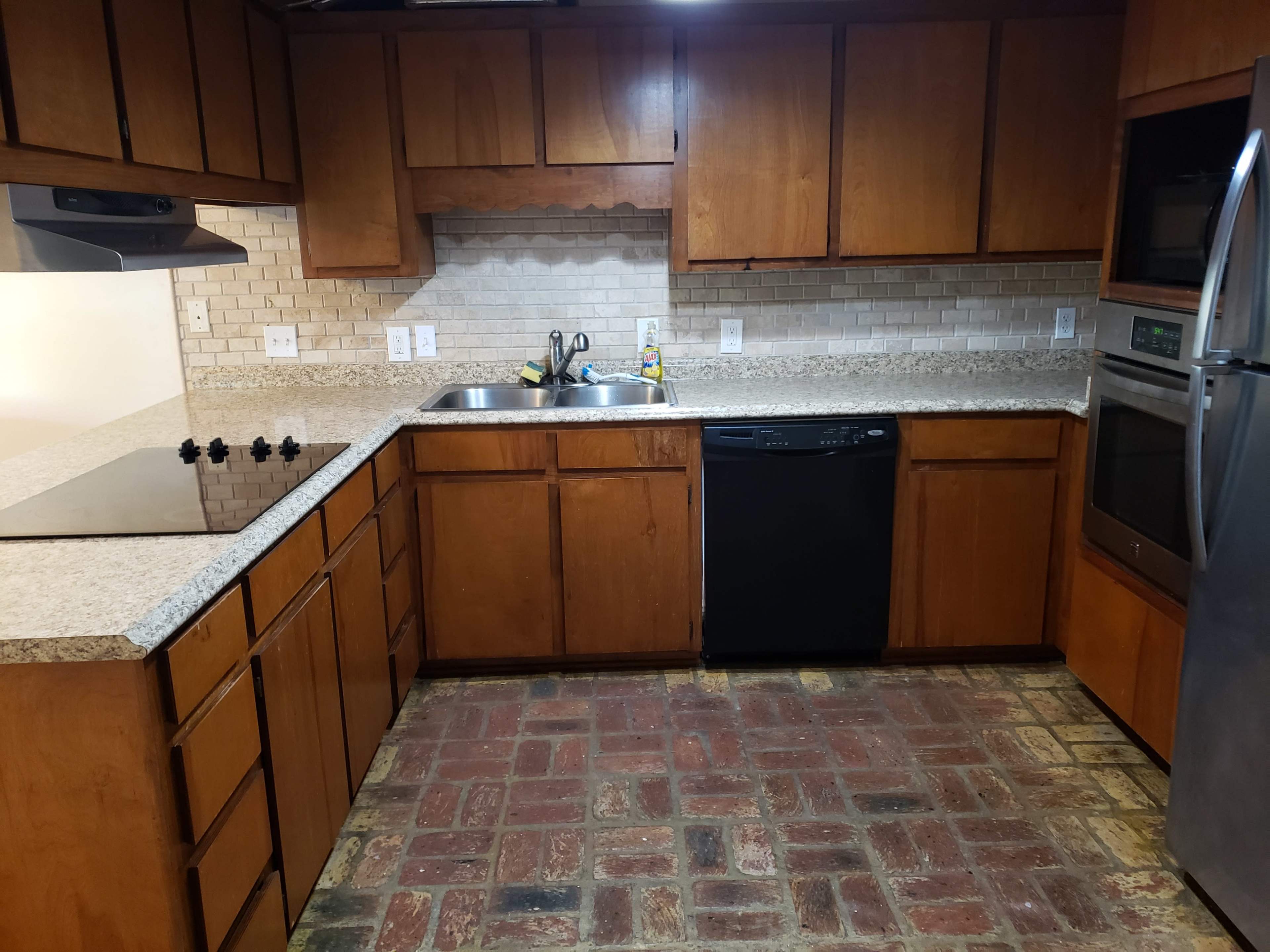 The image shows a kitchen with wooden cabinets, a granite countertop, a sink, a dishwasher, and a brick-patterned floor.