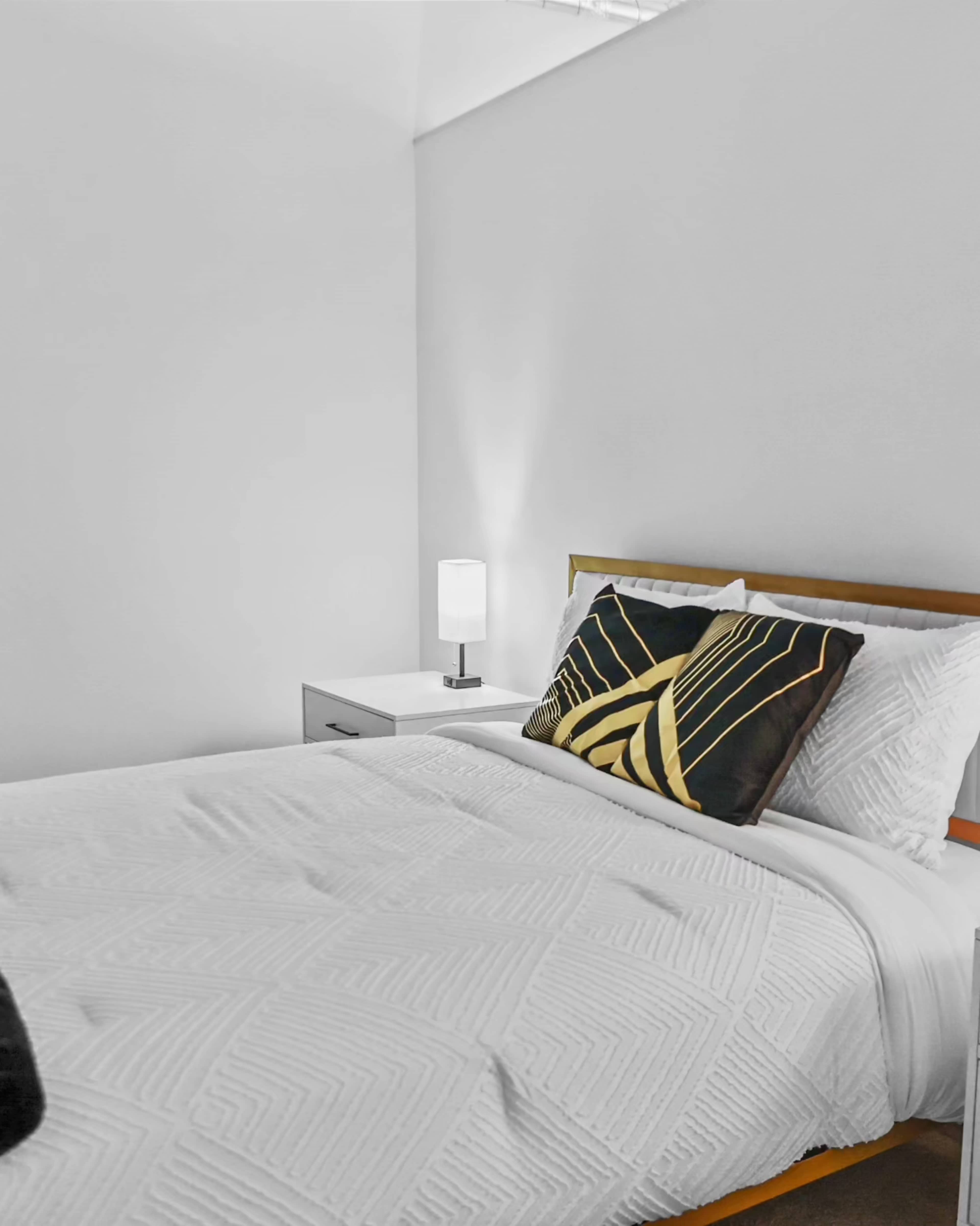 A neatly made bed with a patterned pillow rests against a plain wall, accompanied by a bedside table and a lamp.