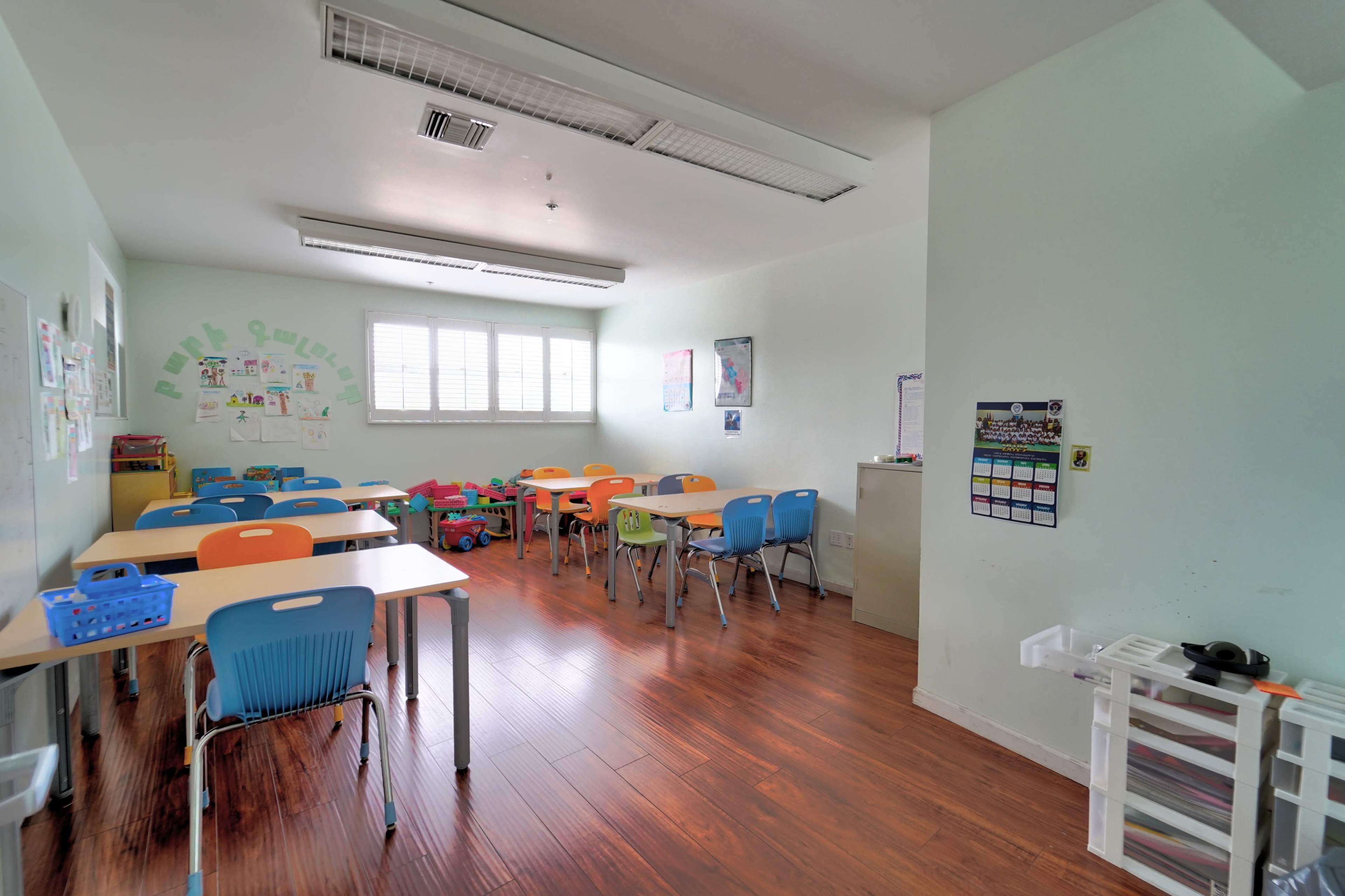 A bright classroom with several tables and chairs arranged, along with children's artwork on the walls and a calendar displayed.