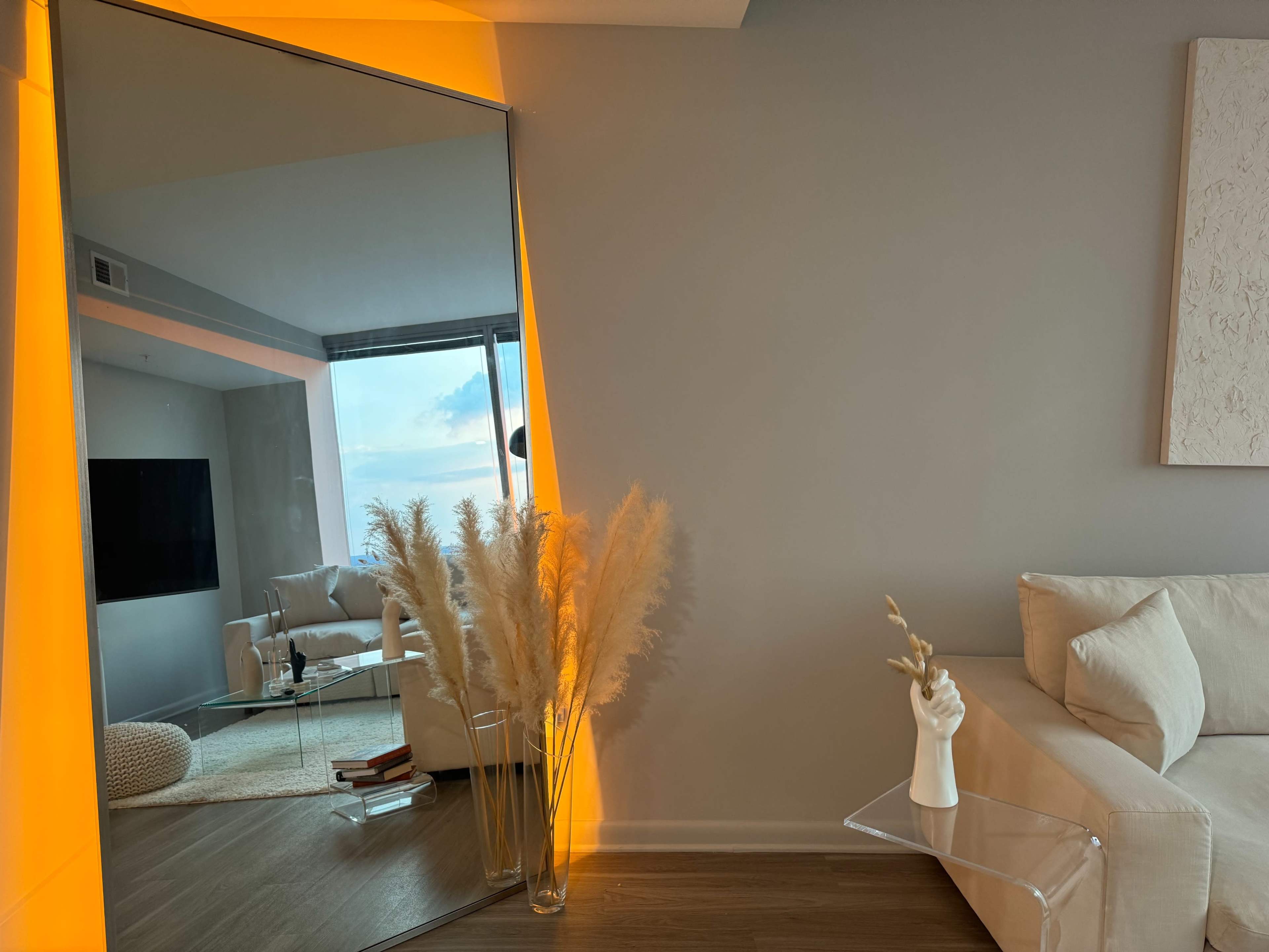 A modern living room featuring a large mirror with orange lighting, a vase of pampas grass, and a clear side table next to a white couch.
