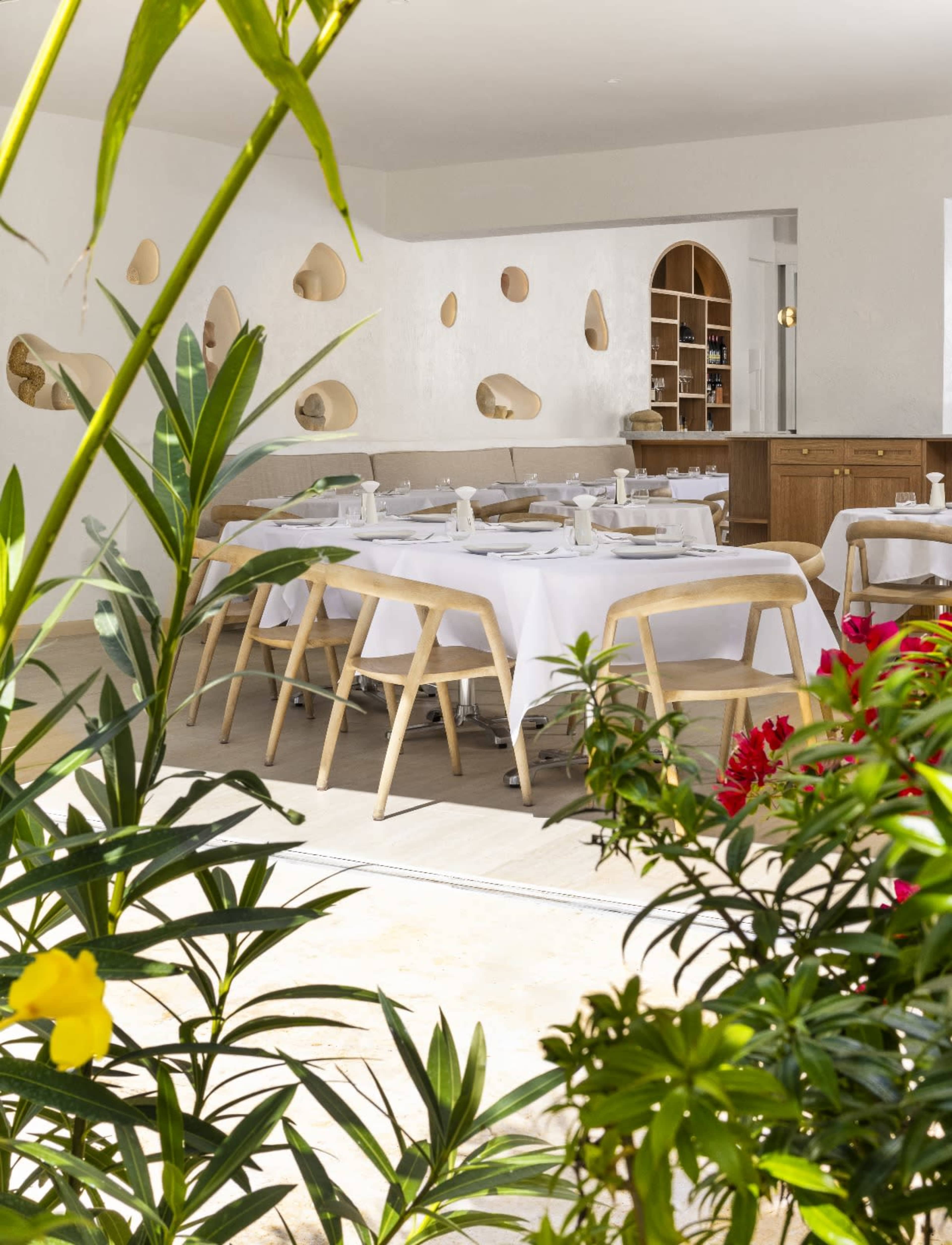 A modern dining area features white tables set neatly with cutlery, surrounded by greenery and textured wall cutouts.