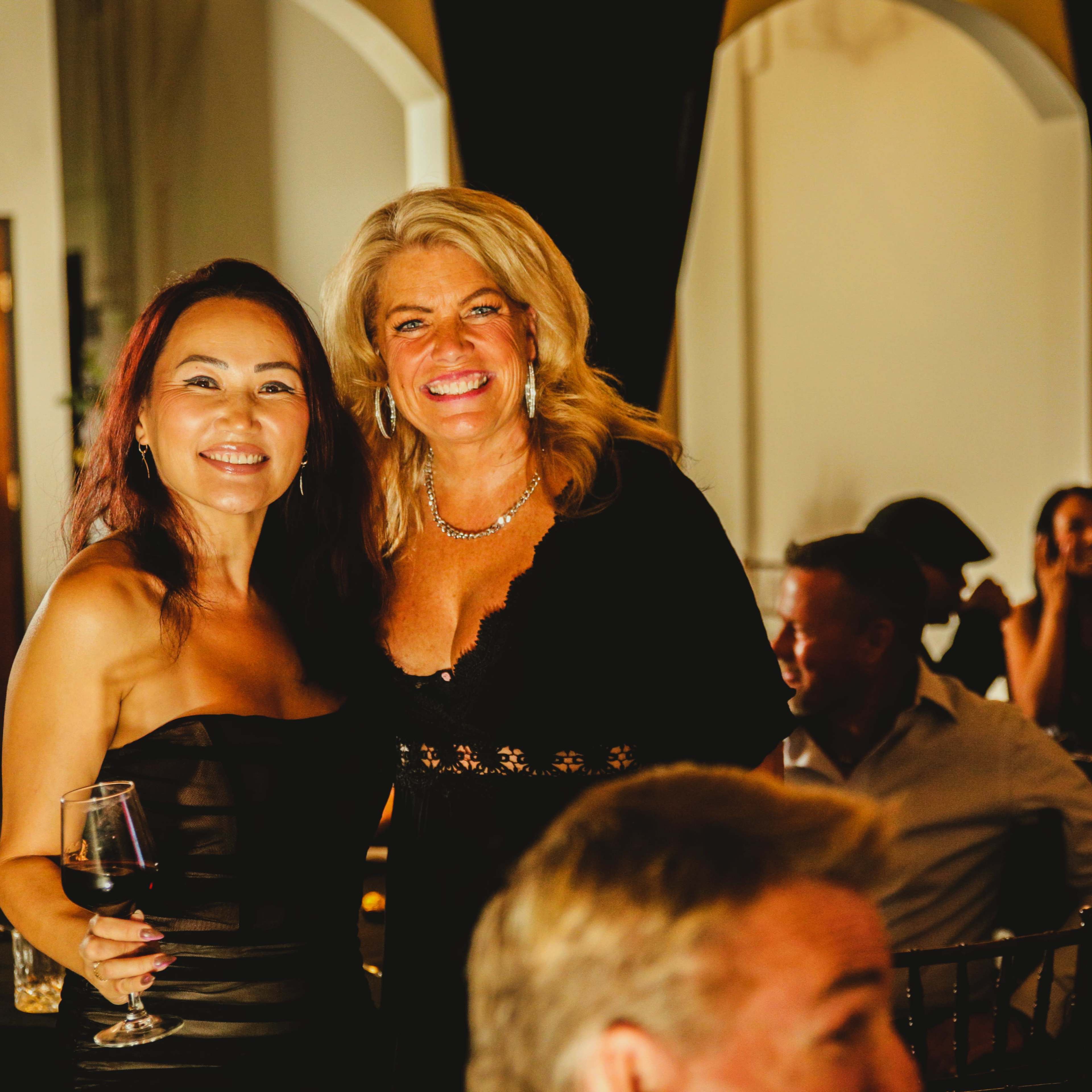 Two women smile and pose for a photo at a social event, with a glass in hand and people mingling in the background.