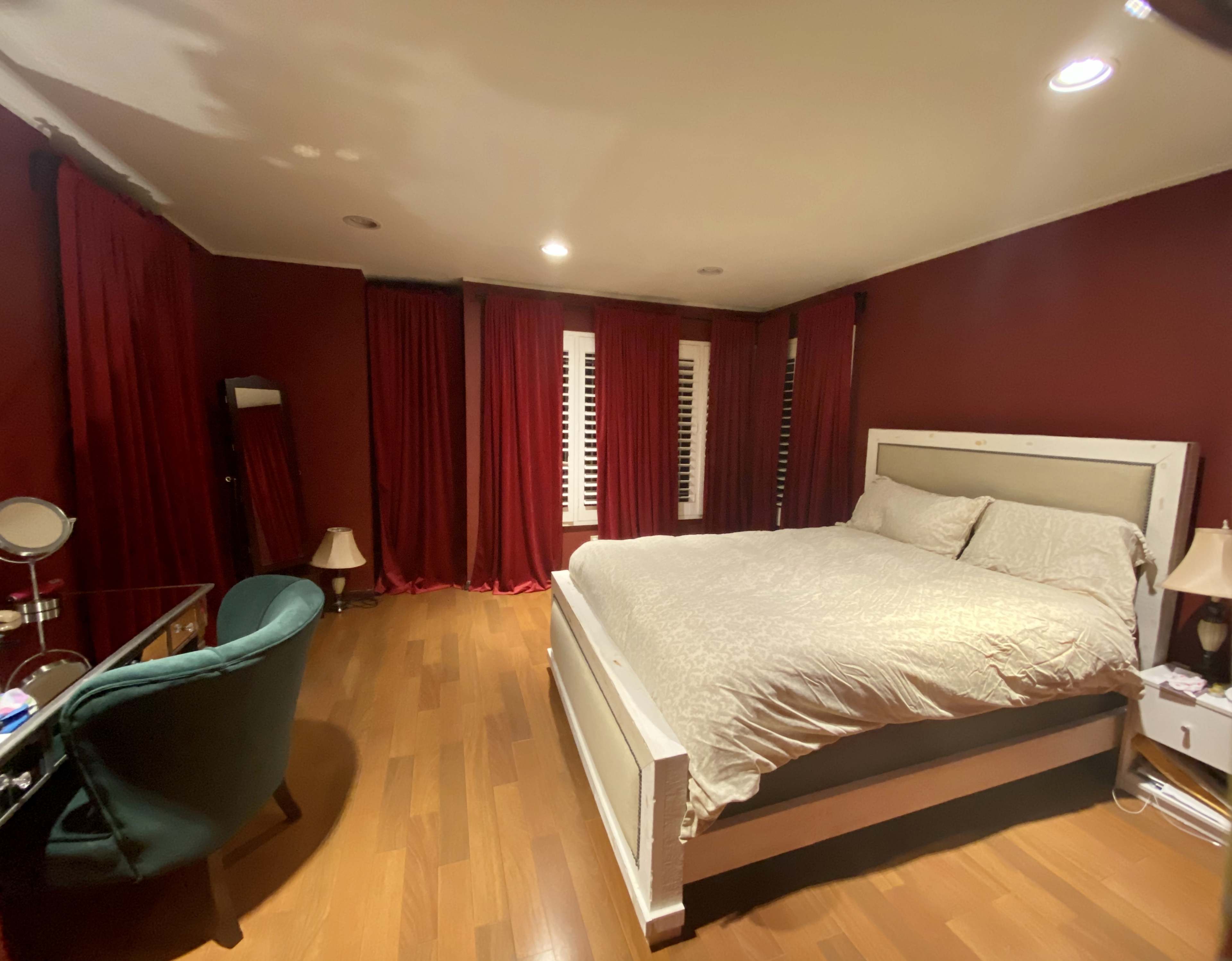 A bedroom features a large bed with a light-colored comforter, surrounded by dark red curtains and hardwood flooring.