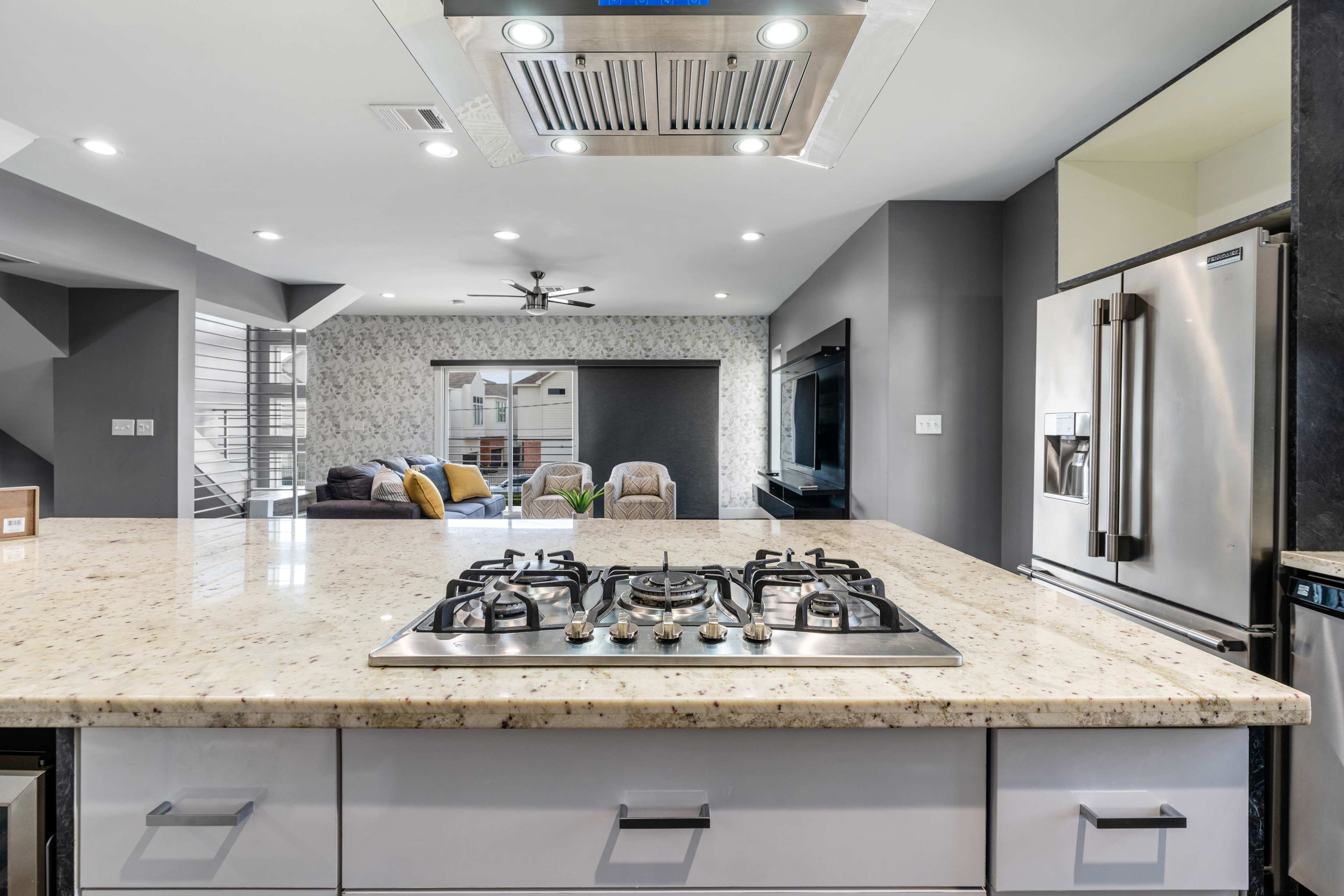 A modern kitchen featuring a granite countertop island with a gas cooktop, stainless steel appliances, and an open living area in the background.