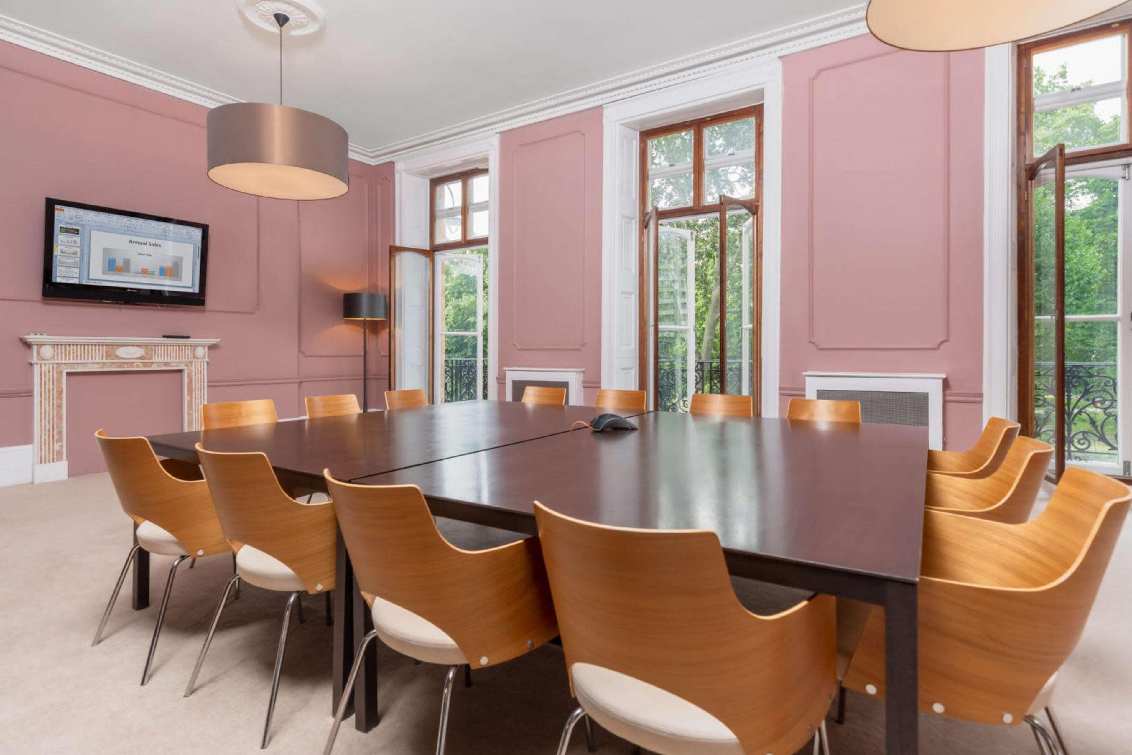 A conference room features a large wooden table surrounded by twelve wooden chairs, with large windows and a wall-mounted screen displaying a presentation.