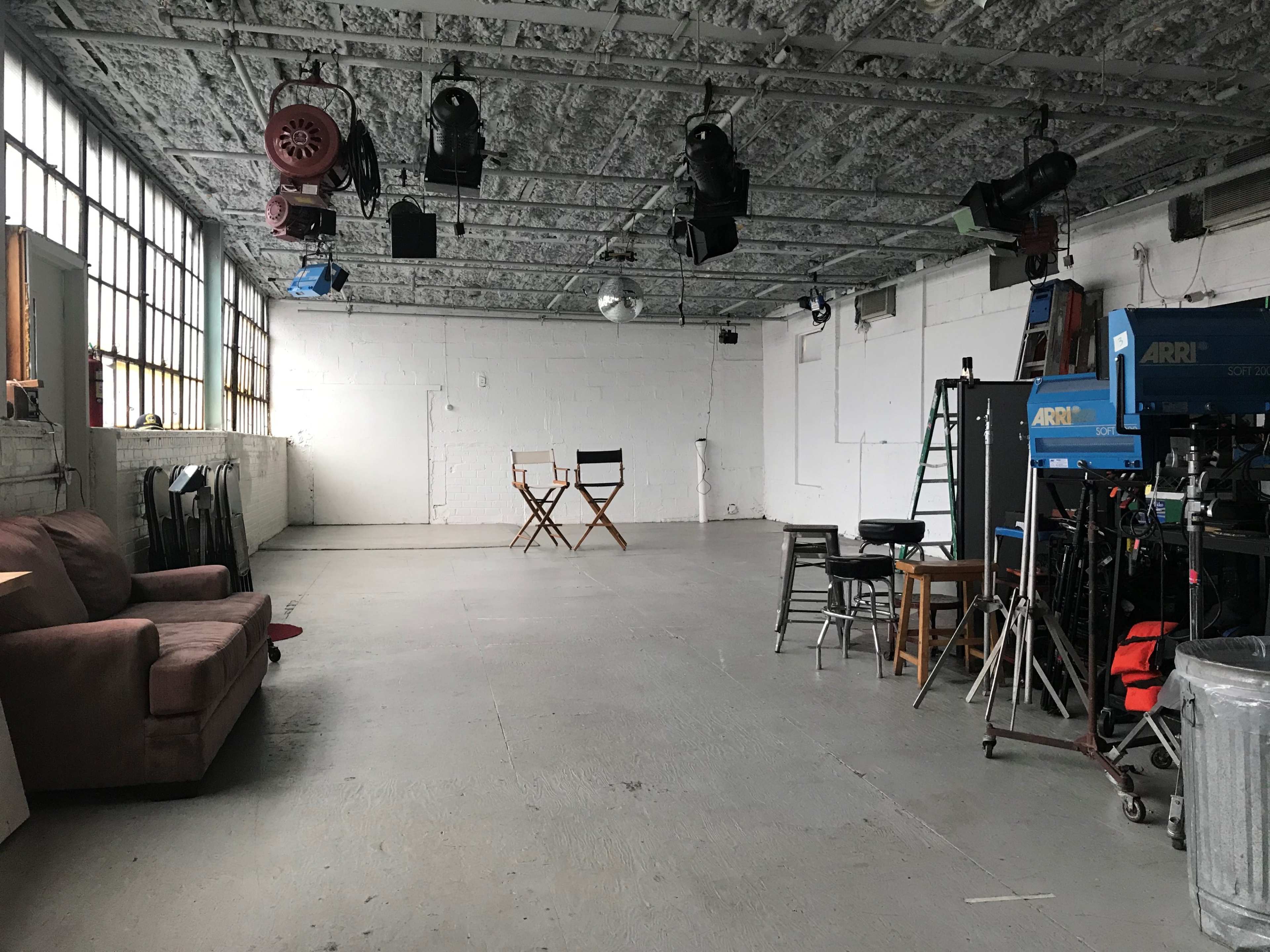 An empty studio space with a sofa, director's chairs, and various lighting and filming equipment.