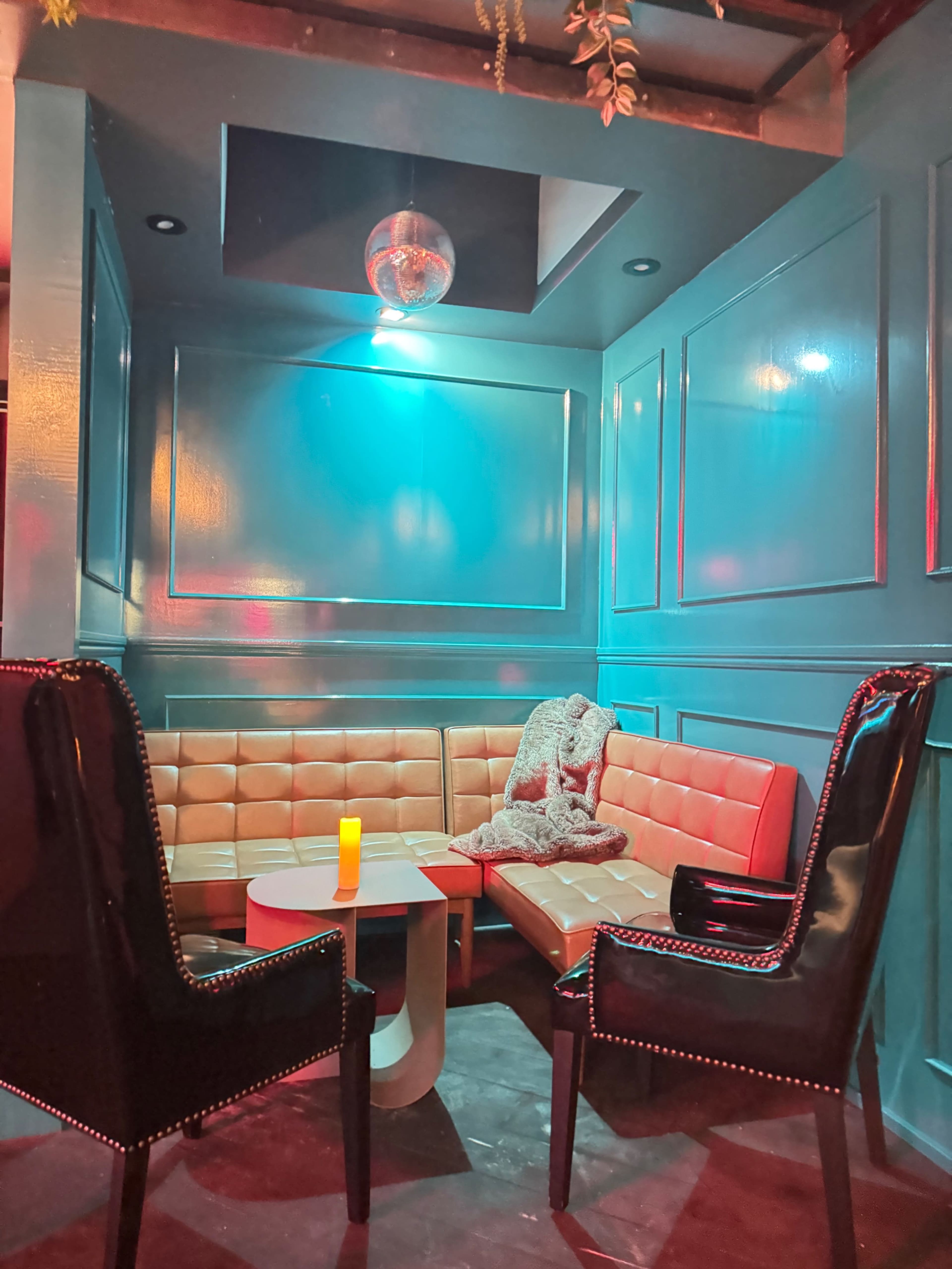 A cozy corner seating area features orange and cream-colored upholstered benches, black chairs, and a small white table, illuminated by soft, colored lights and a single candle.